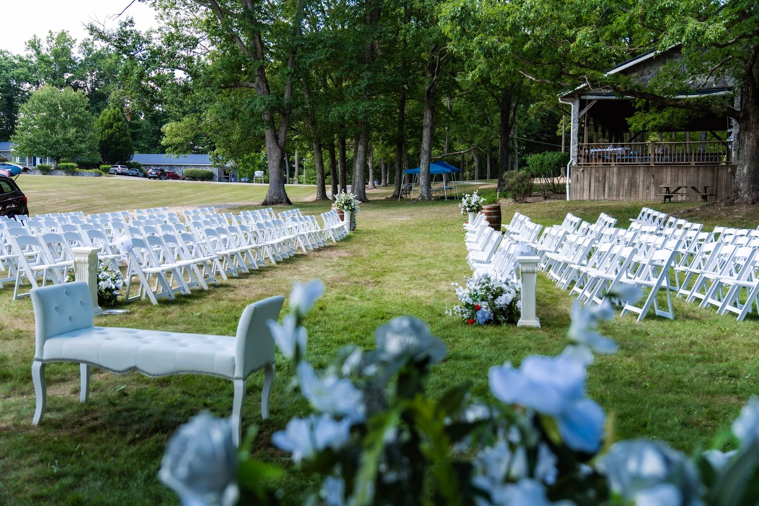 Wedding venue setup with white chairs and blue floral decorations in a wooded outdoor setting.