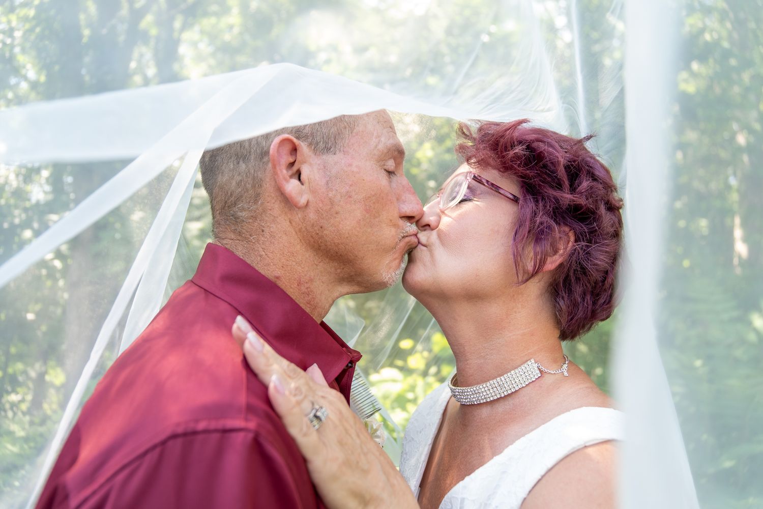 A romantic wedding kiss shared between a couple outdoors with a white veil flowing in sunlit woodland scenery.