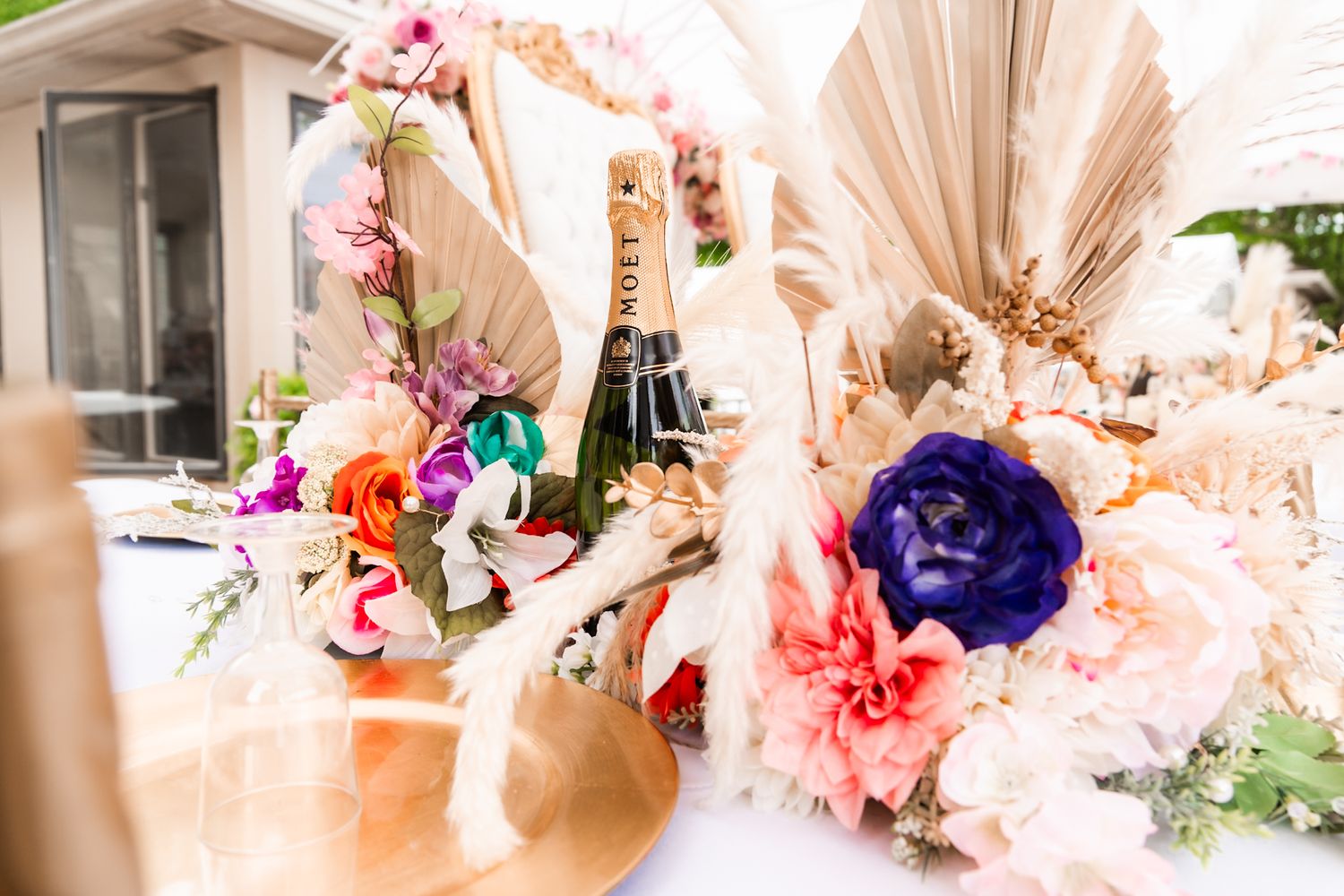 Elegant wedding reception table with champagne bottle, dried palm leaves and colorful floral arrangements.