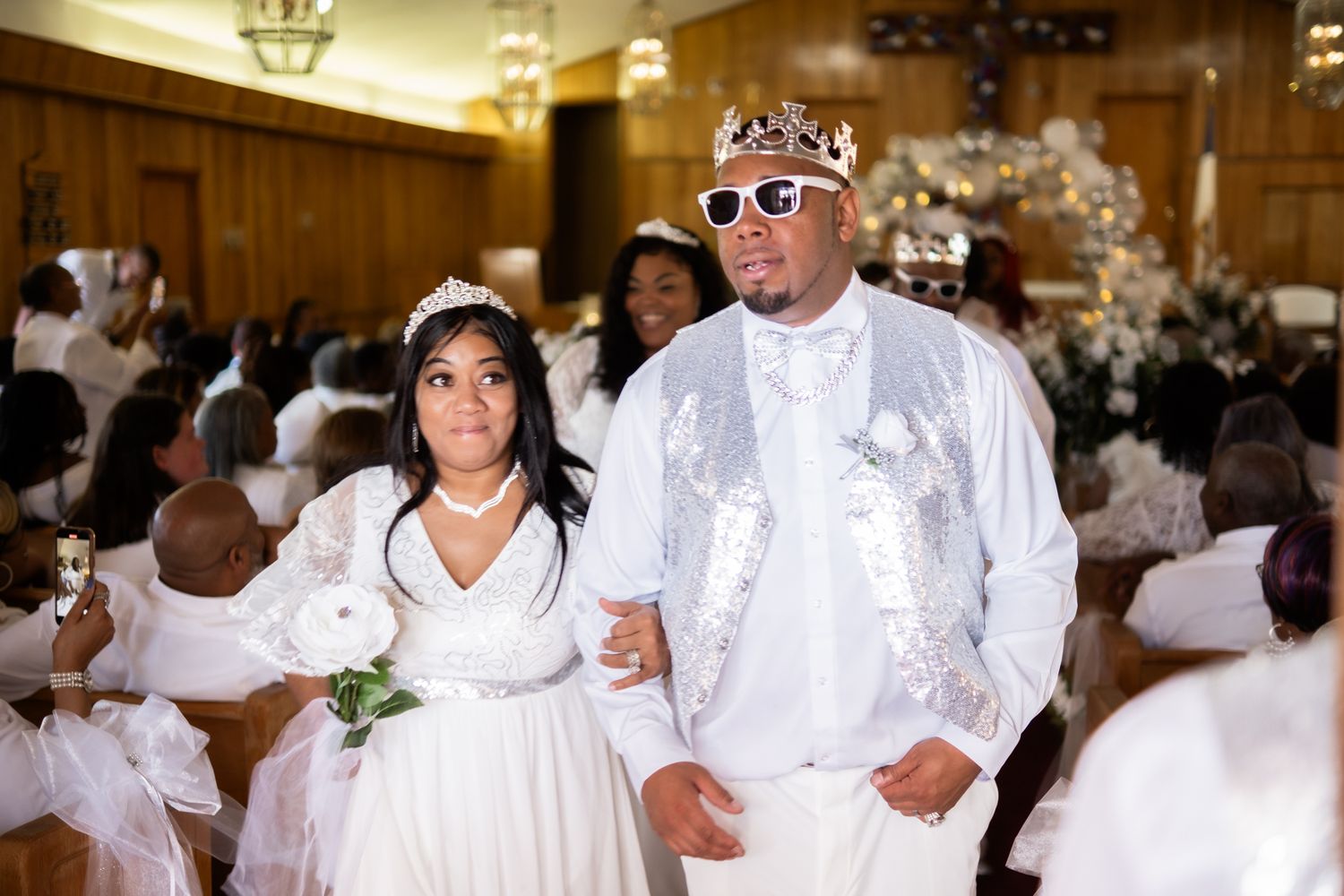 Wedding celebration scene with couple in white attire and crowns during an indoor reception ceremony.