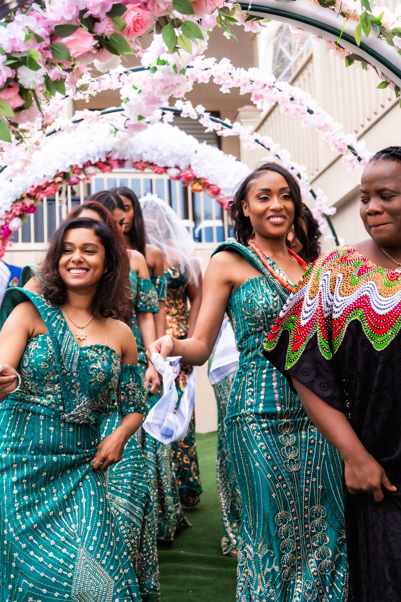 Bridesmaids in sparkling turquoise dresses celebrate under pink floral arch at wedding.