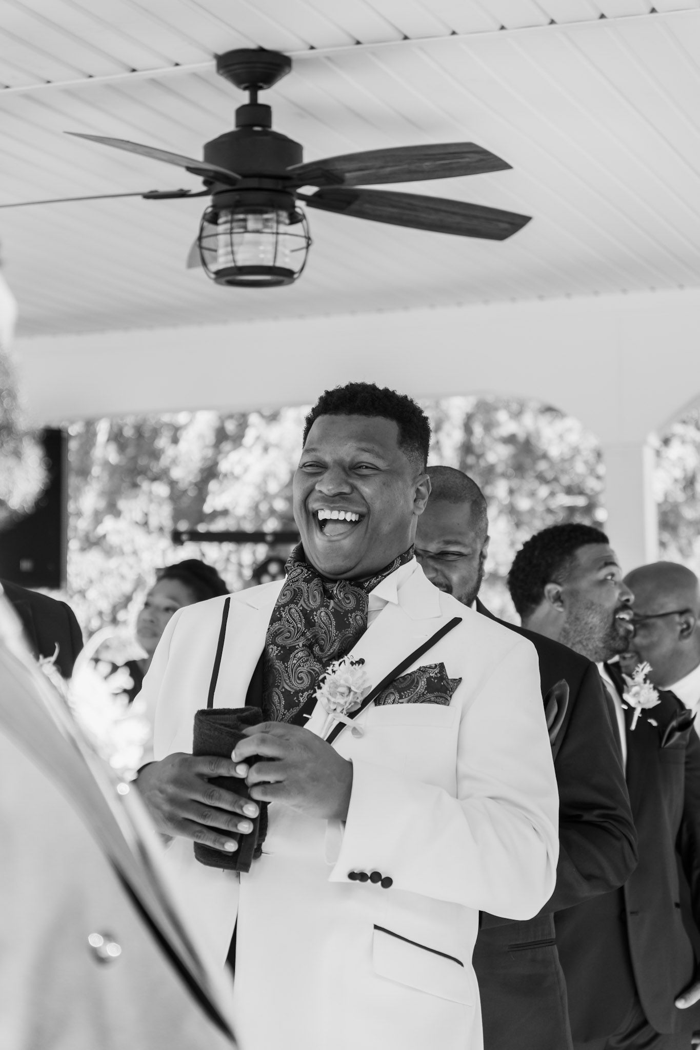 Wedding guest in white suit jacket laughing during outdoor reception.