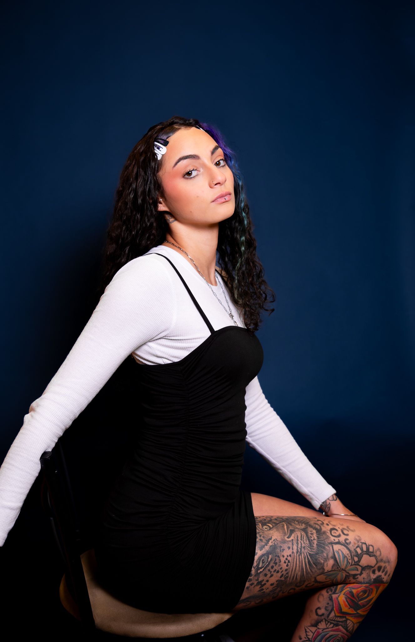 Studio portrait of a young model in a black dress and white long sleeve top showing leg tattoos against dark background.