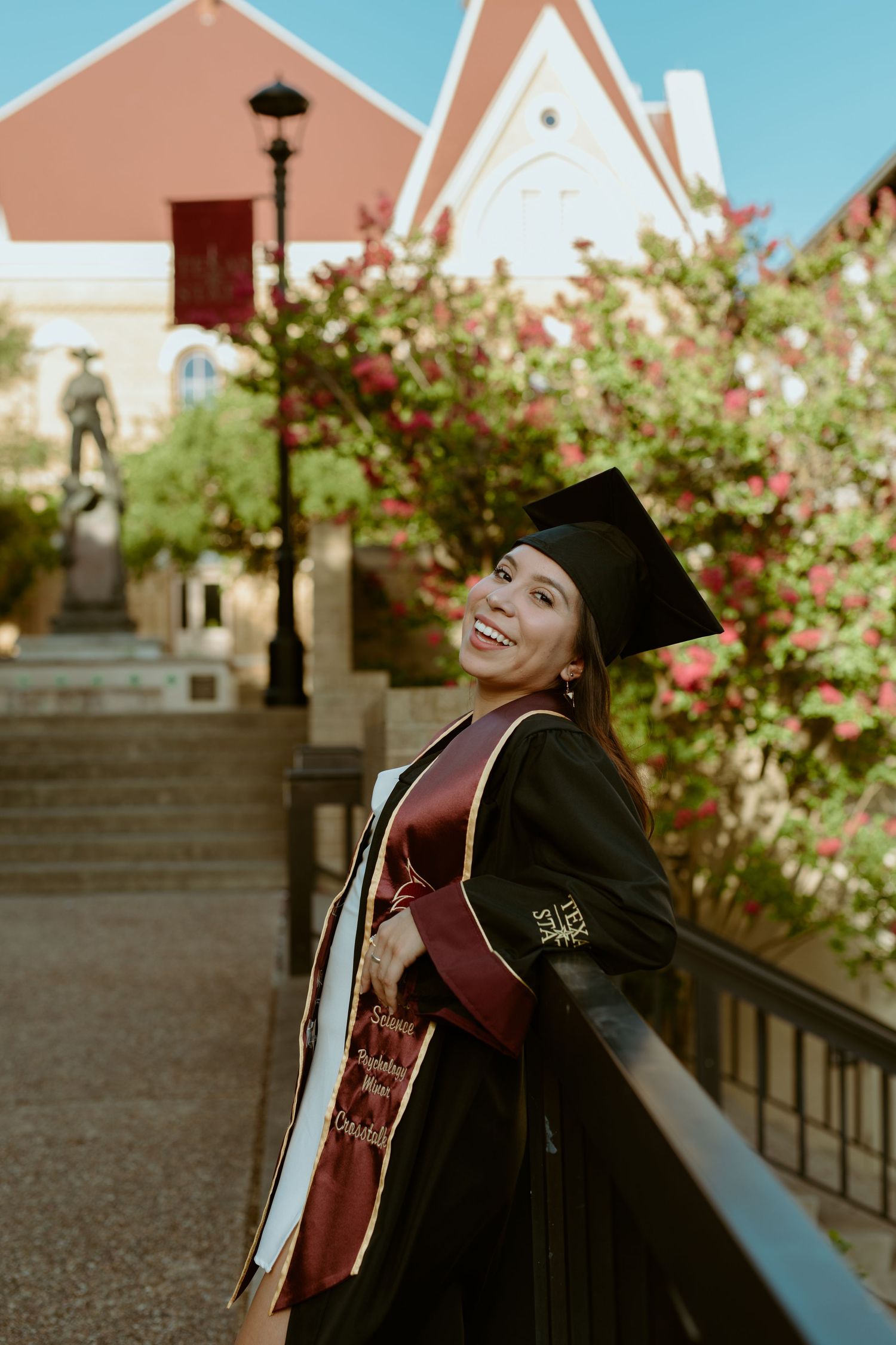 A Texas State University graduate smiling in her cap and gown while leaning against a black railing on campus in San Marcos, Texas.
