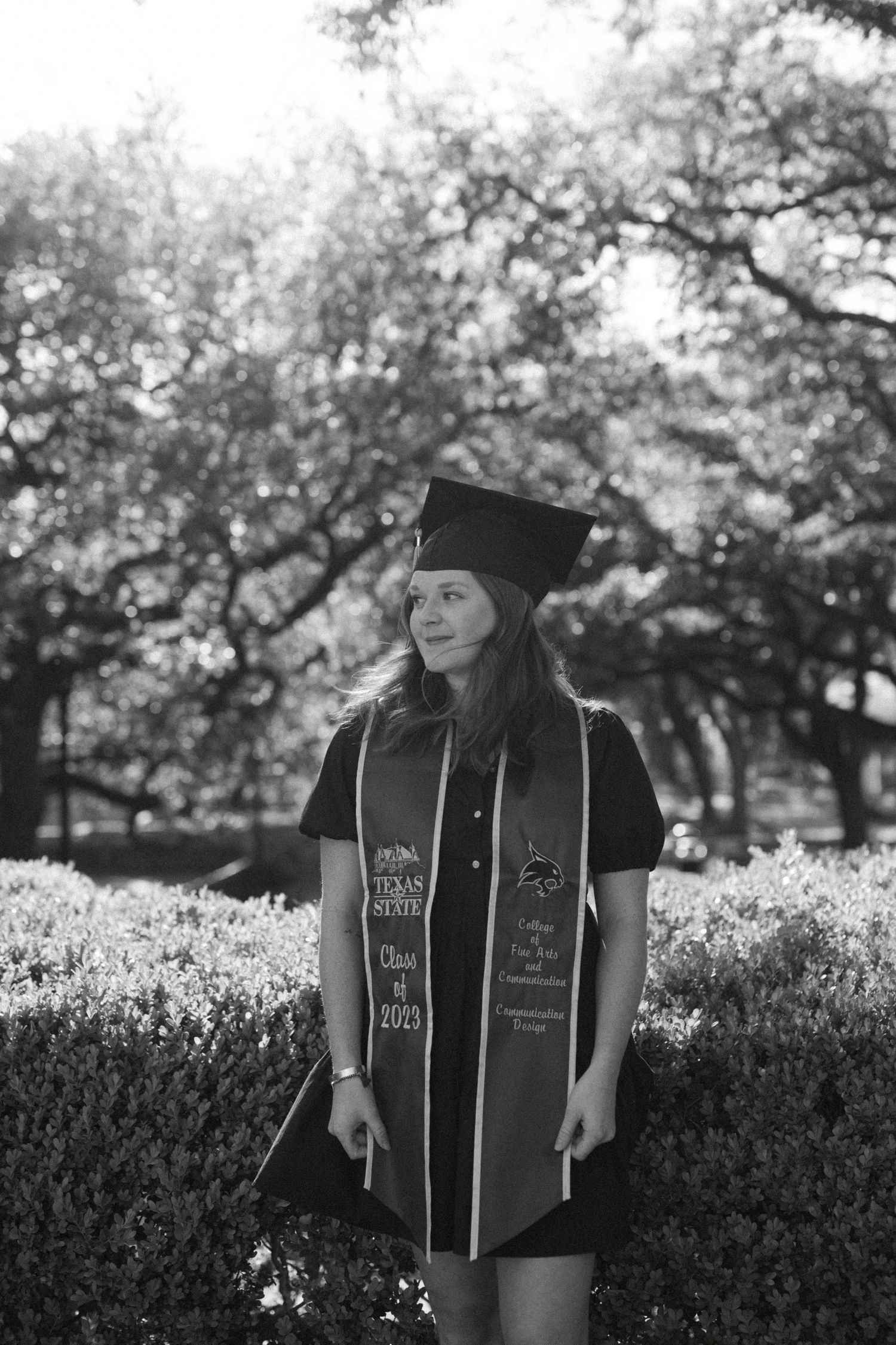 Black and white graduation photography of a Texas State University senior in a cap and gown standing in front of lush trees in San Marcos TX
