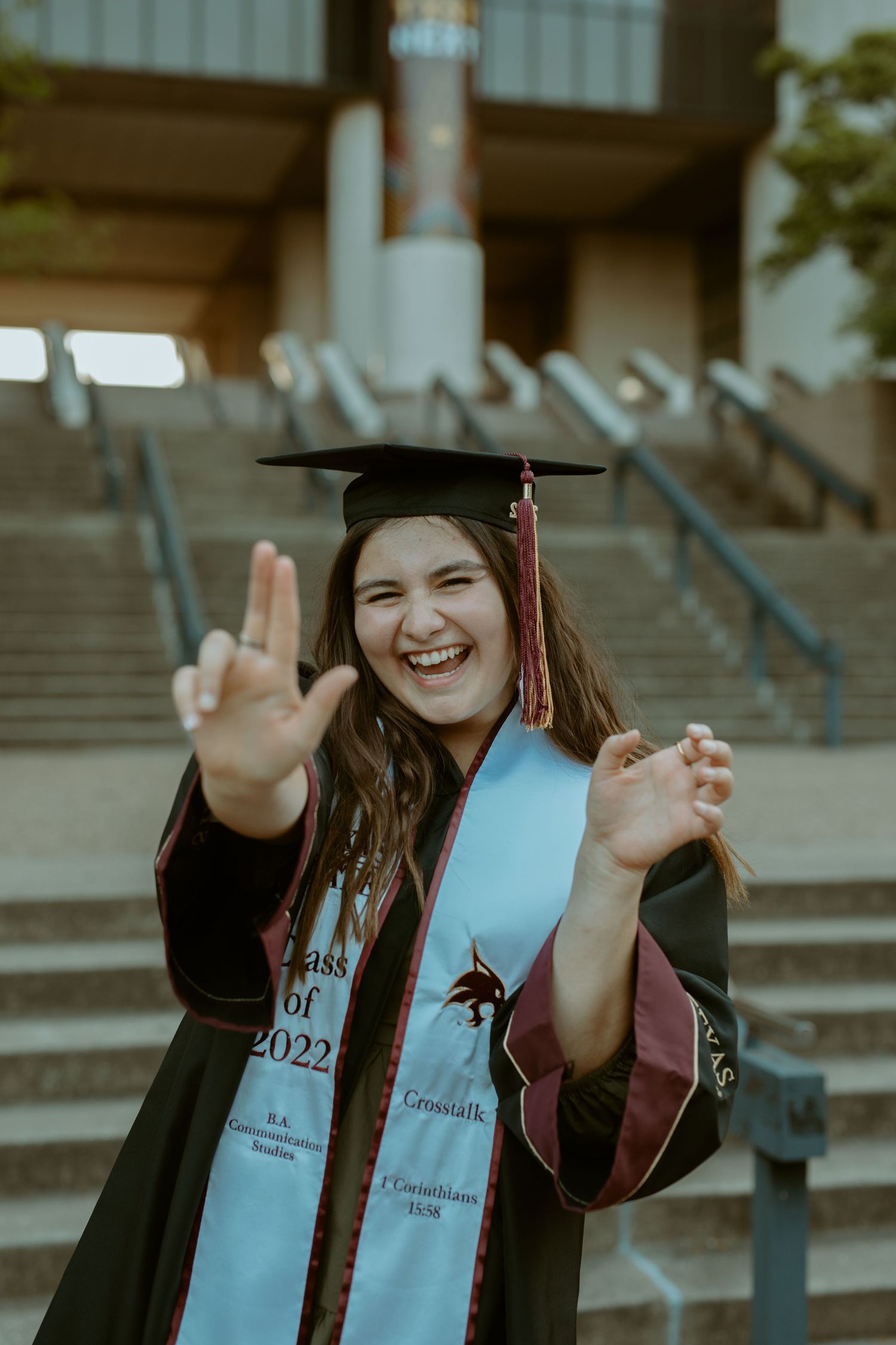 A smiling Texas State University graduate in a cap and gown flashing the 