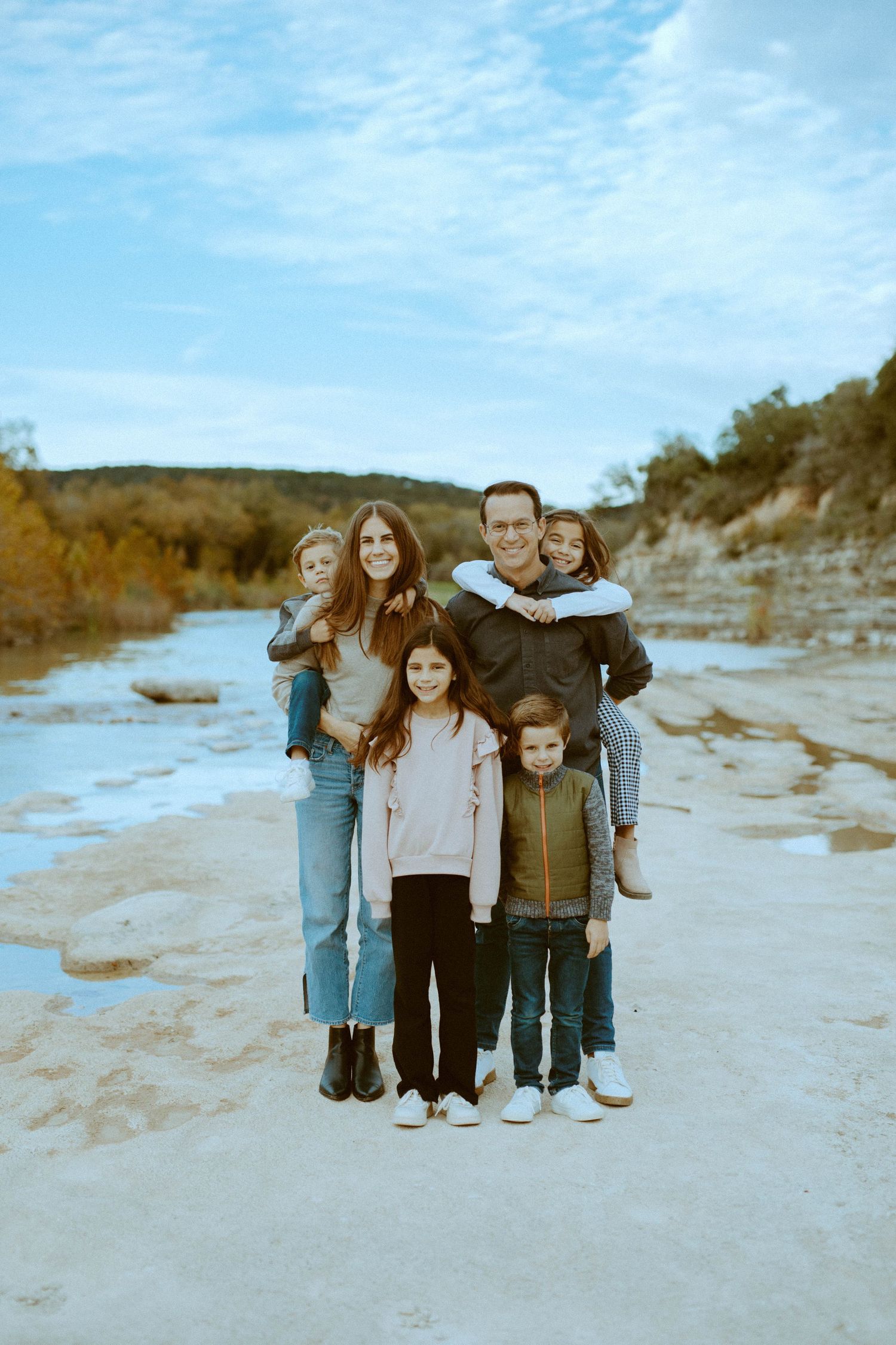 A smiling family of six posing together on the rocky banks of the Blanco River during a photo shoot in Wimberley, Texas.