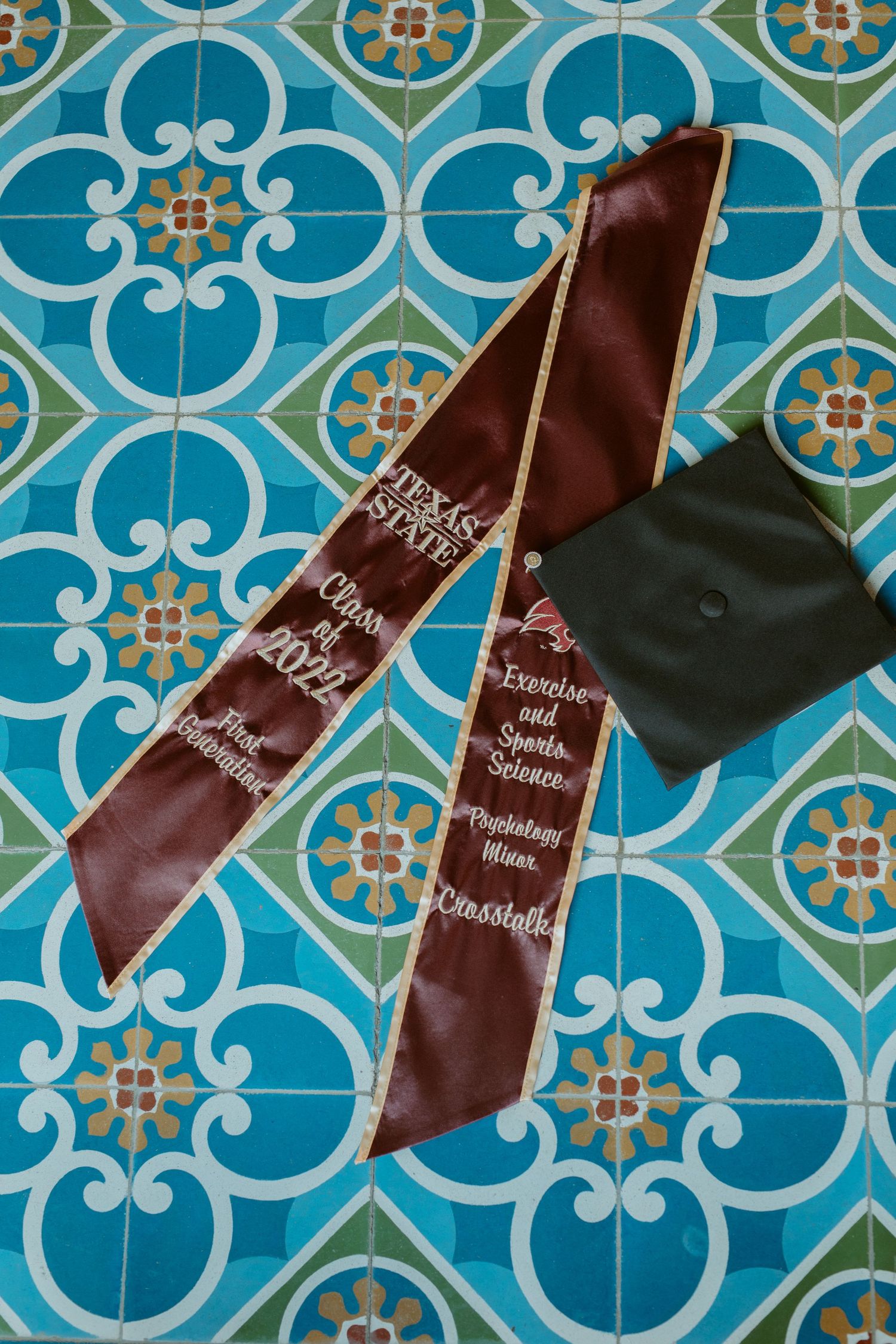 Flat lay photography of a Texas State graduation stole and cap arranged on the iconic blue and yellow patterned tiles at Texas State Univers