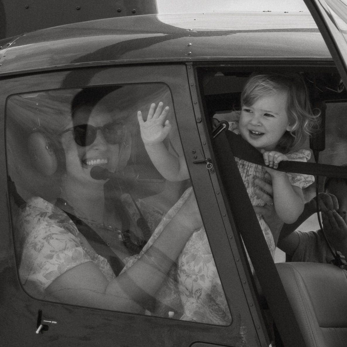 Black and white photo of a smiling mother wearing aviation headsets waving from inside a helicopter next to her excited toddler.