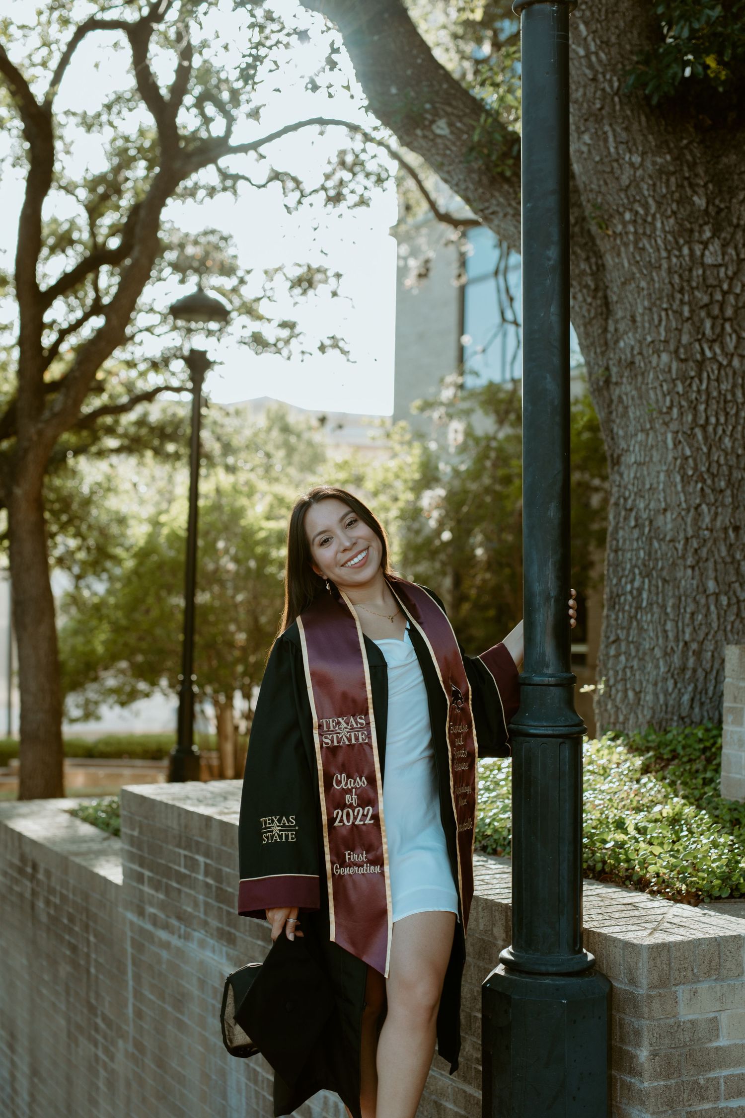 A Texas State University graduate smiling in her cap and gown while standing next to a stone retaining wall under oak trees in San Marcos, T