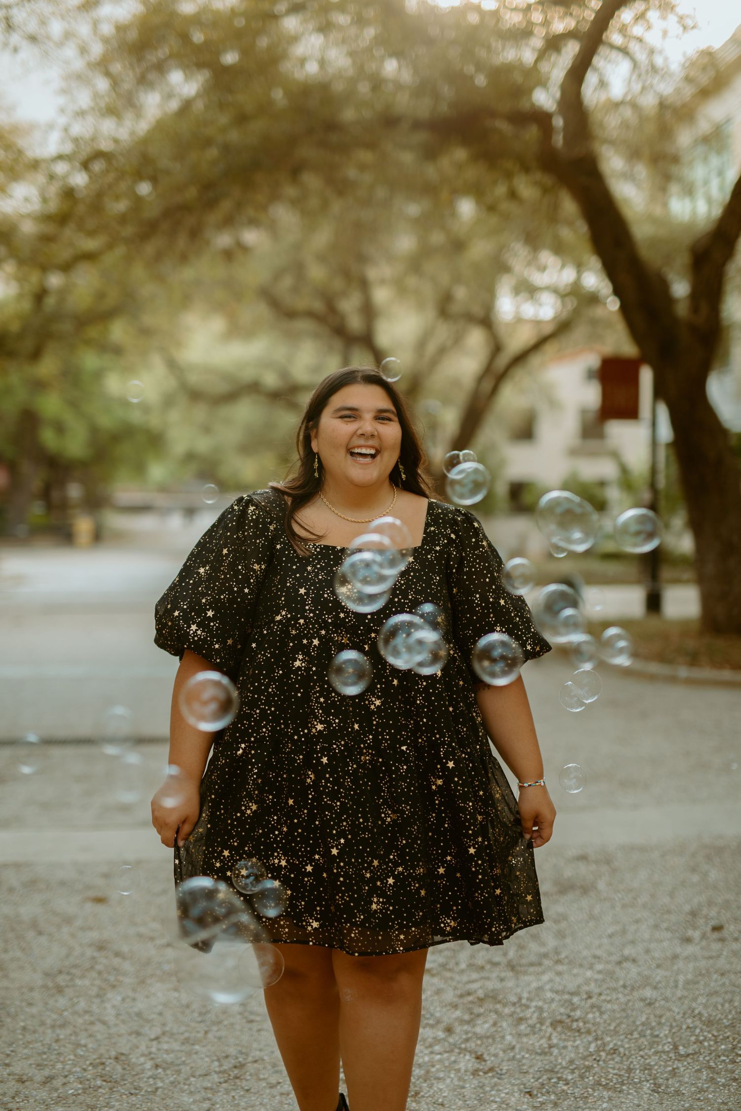 Candid graduation photography of a smiling senior celebrating with bubbles on a tree-lined walkway at Texas State University in San Marcos,
