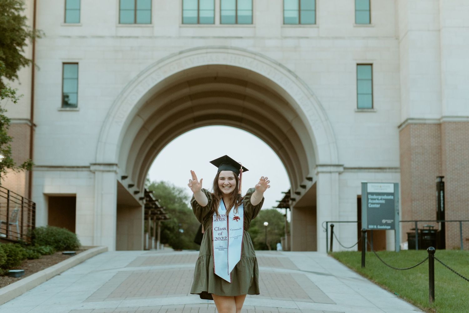 Graduate posing in a cap and gown under a large stone archway during a senior portrait session in San Marcos, TX.