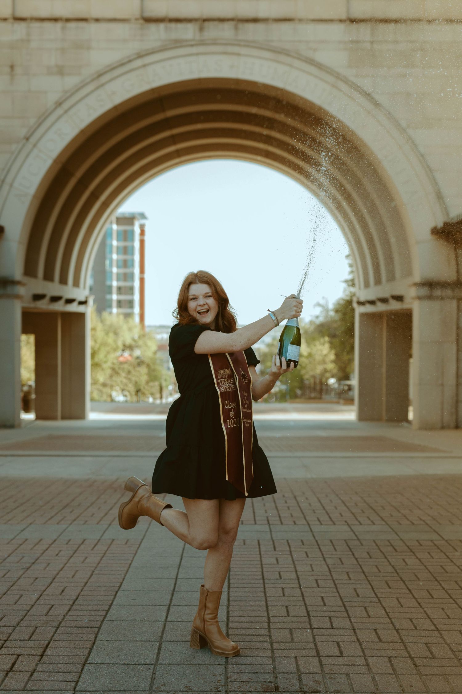 Texas State University graduate spraying champagne under the campus archway during a senior portrait session in San Marcos, TX.