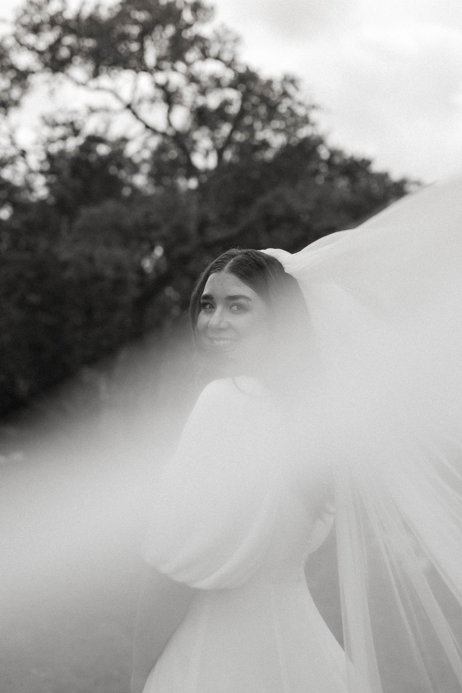 Creative black and white bridal portrait in Wimberley, TX, featuring a bride smiling over her shoulder with her wedding veil catching the wi