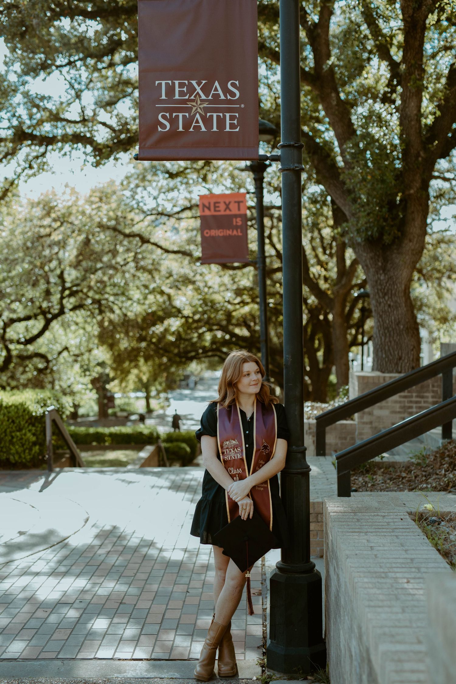 A Texas State University graduate wearing a maroon stole stands by a lamp post with university banners on the San Marcos campus.