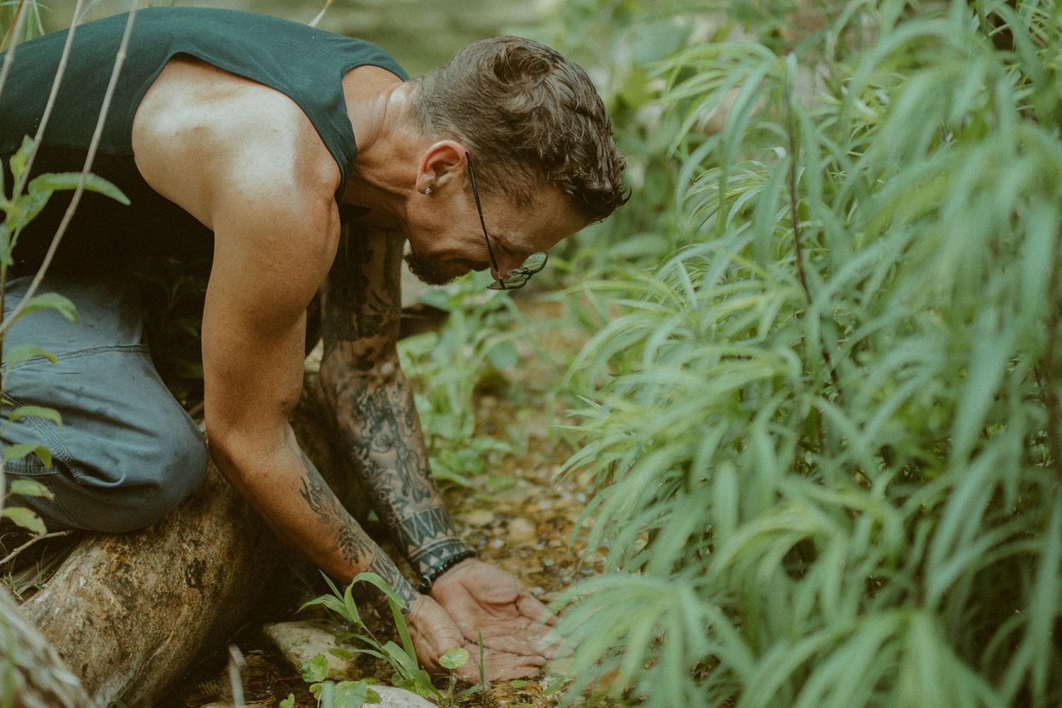 A tattooed man in a tank top leaning down over a log to scoop fresh water with his hands in a lush, green forest.
