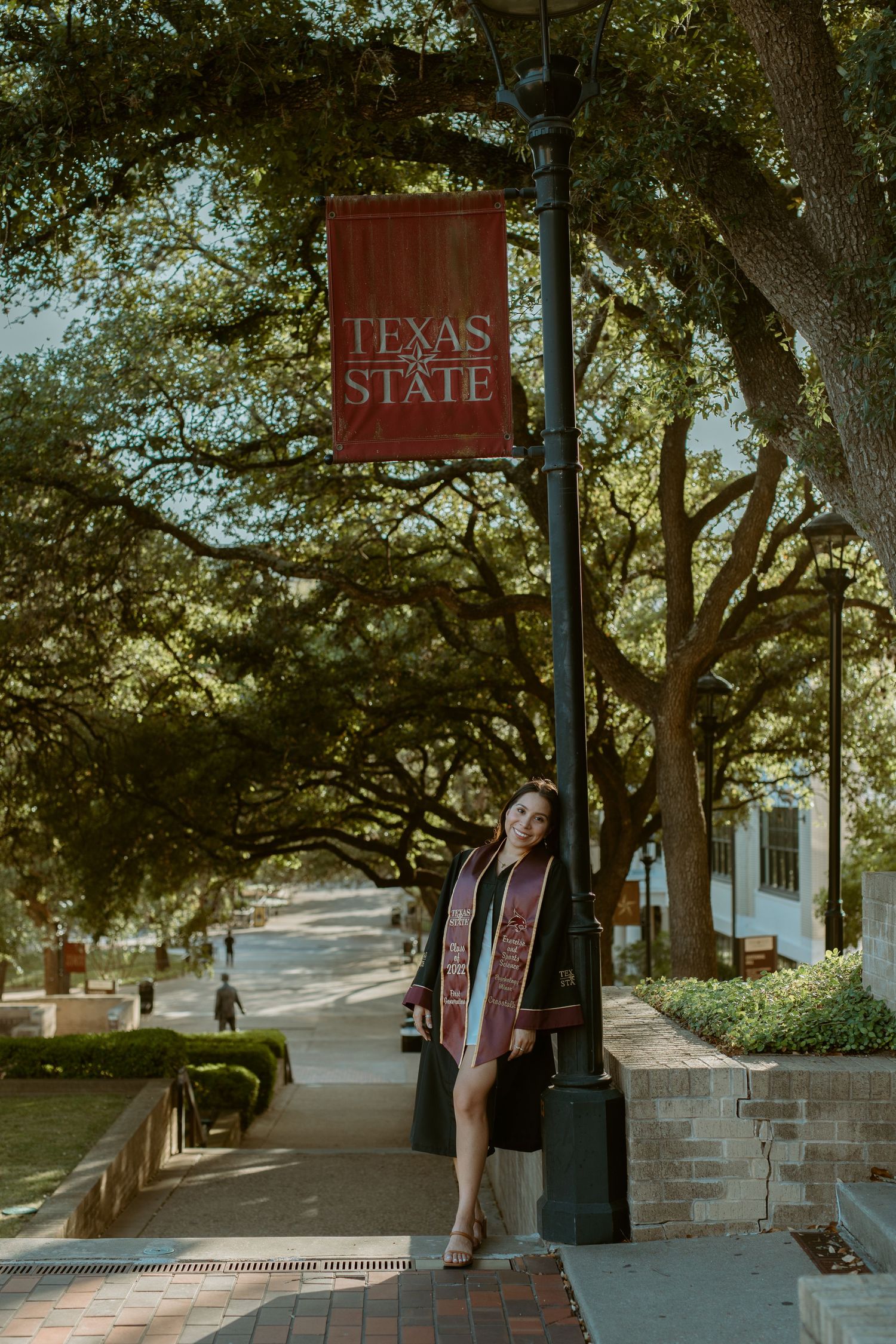Wide-angle graduation portrait of a senior leaning against a lamp post beneath a maroon Texas State banner on campus in San Marcos, Texas.