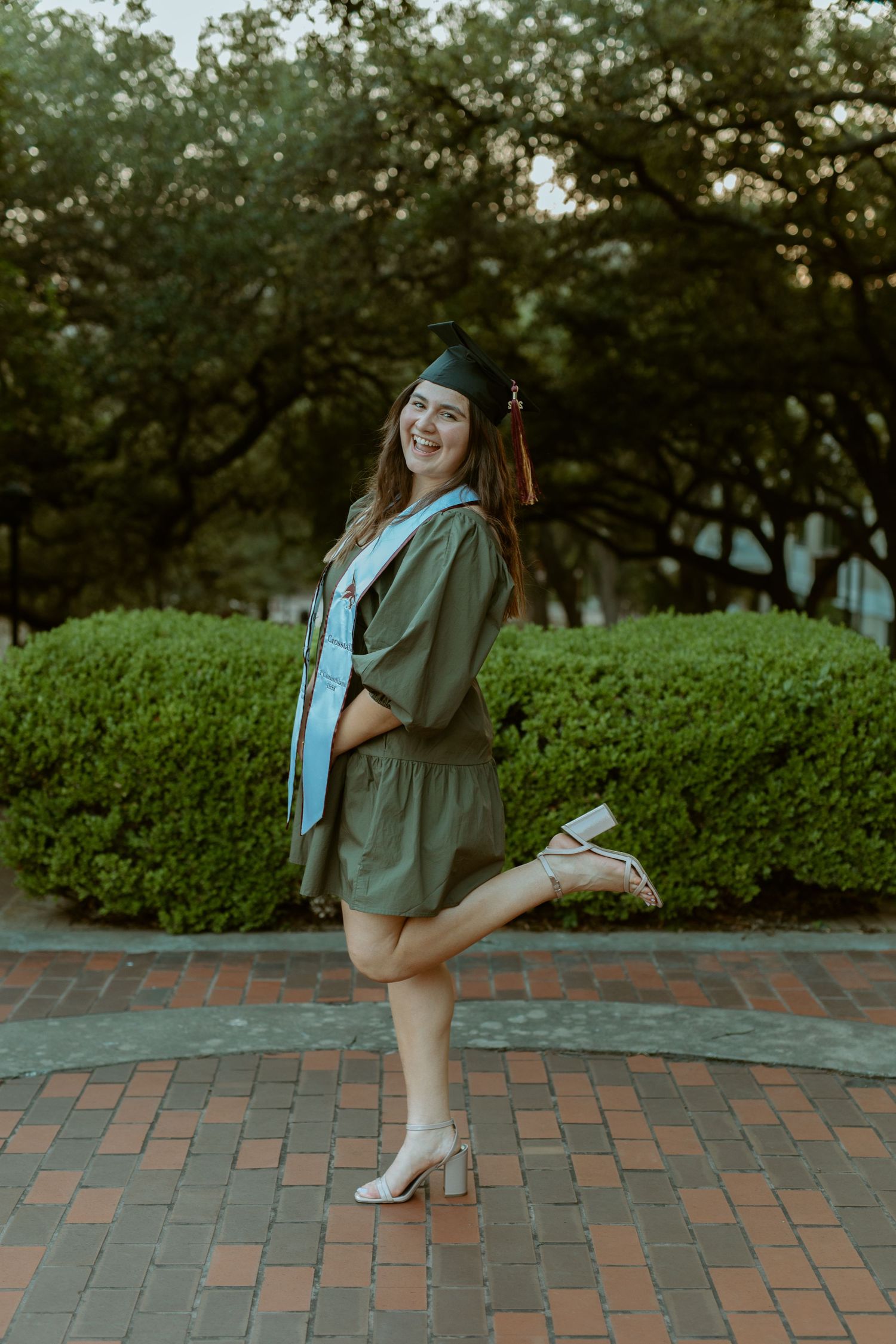 A Texas State University graduate in a cap and gown posing playfully on a brick pathway on campus in San Marcos, Texas.