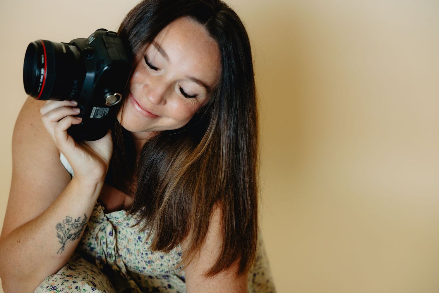 A young woman with long brown hair smiles while holding a Canon DSLR camera against a beige background.