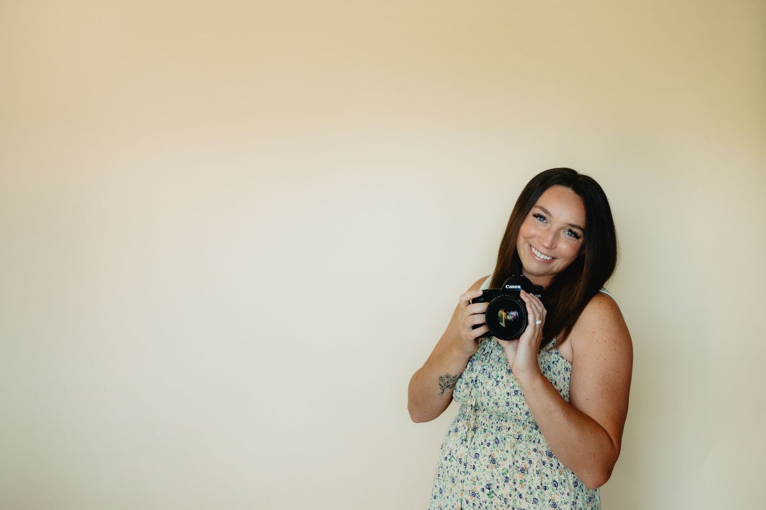 Smiling woman holding a camera, wearing a floral dress, standing against a light beige wall.
