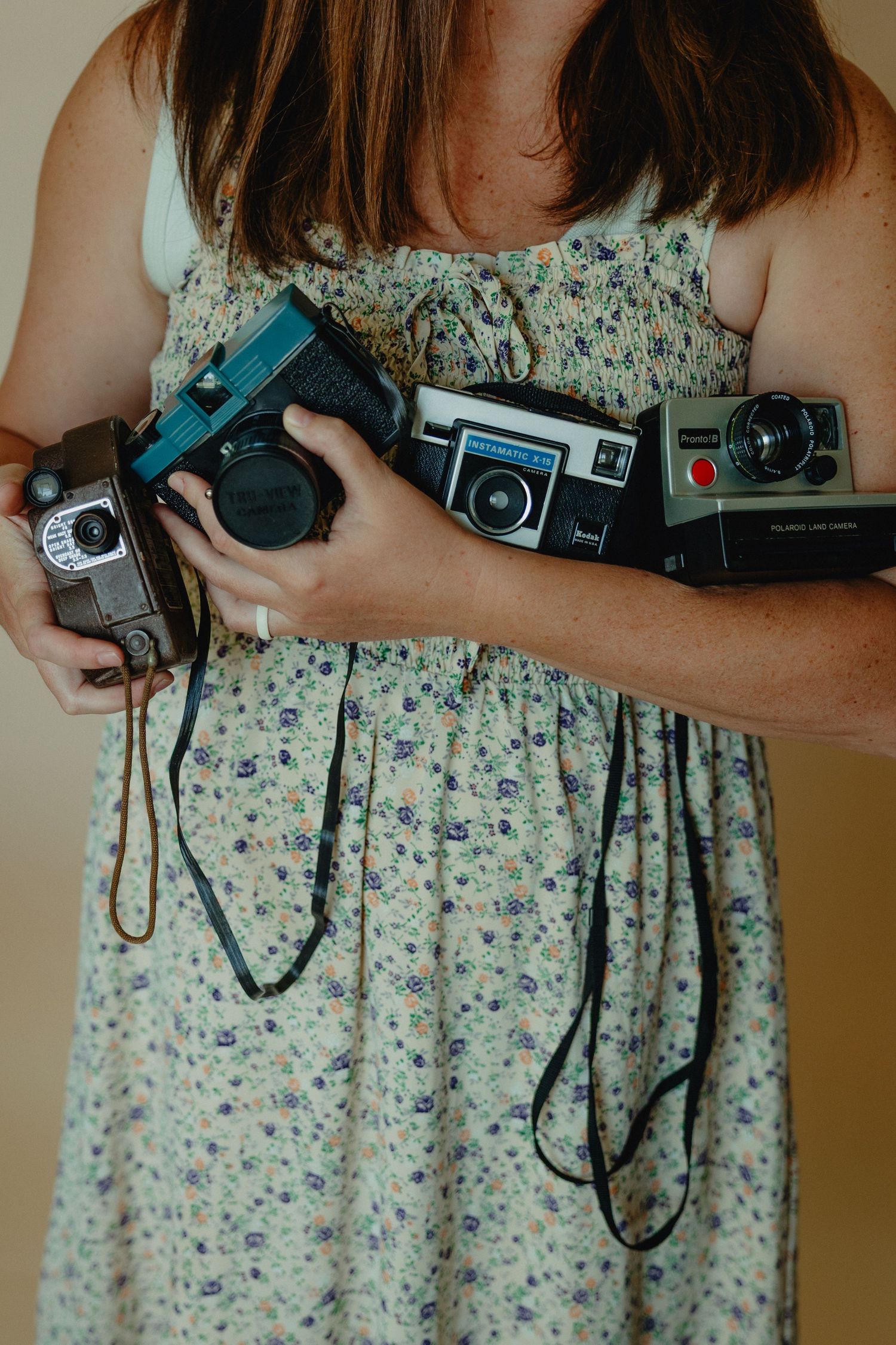 Woman in floral dress holding multiple vintage cameras including Polaroid and film cameras.