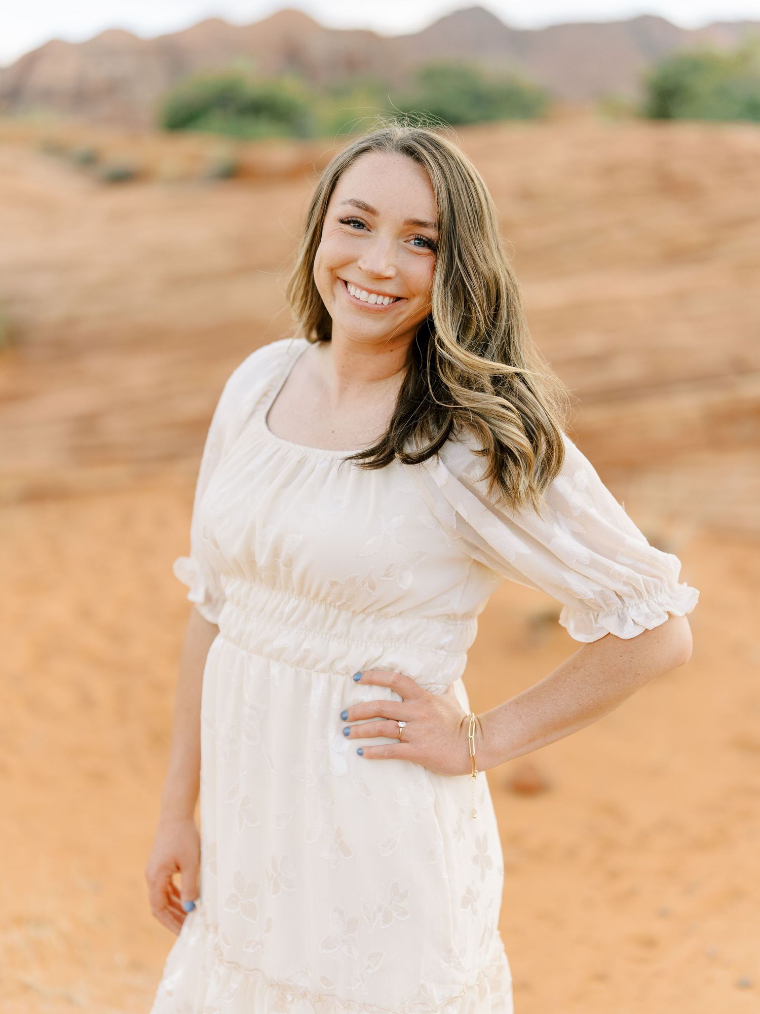 A person in a white dress with long wavy hair poses outdoors in a desert landscape.