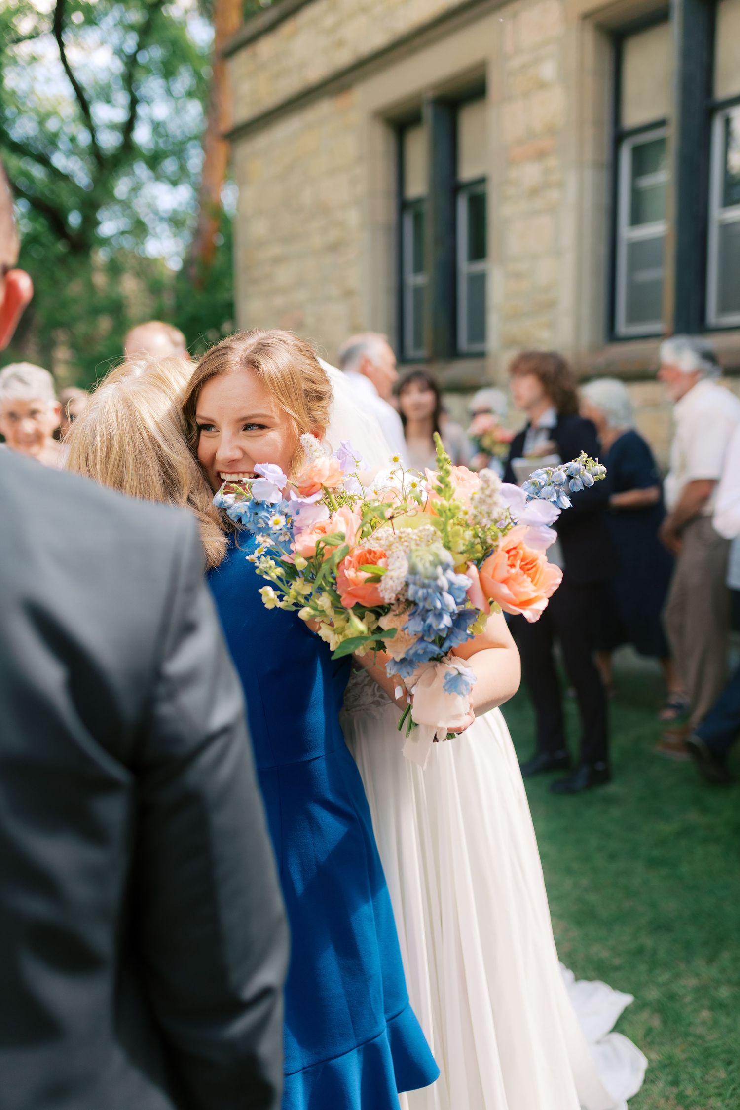 Smiling bride in white gown hugging guest while holding colorful peach and blue bridal bouquet.