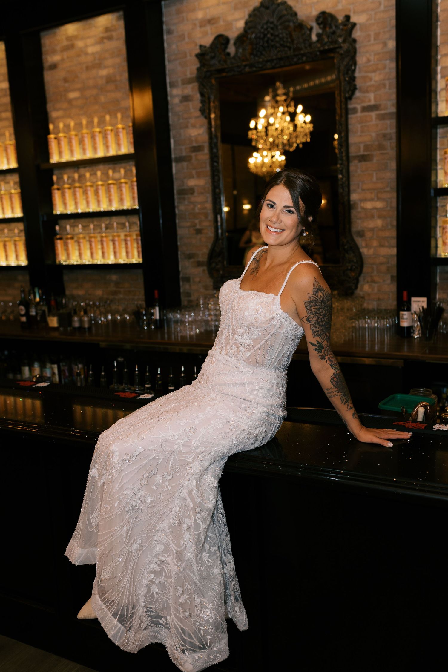 Tattooed bride in white lace gown sitting on a bar counter, smiling, with elegant chandelier and brick wall backdrop.