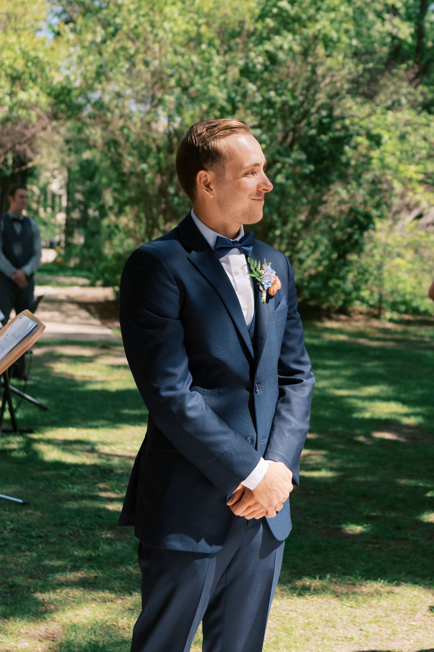 Groom in navy blue suit with bow tie and boutonniere stands outdoors at garden wedding ceremony.