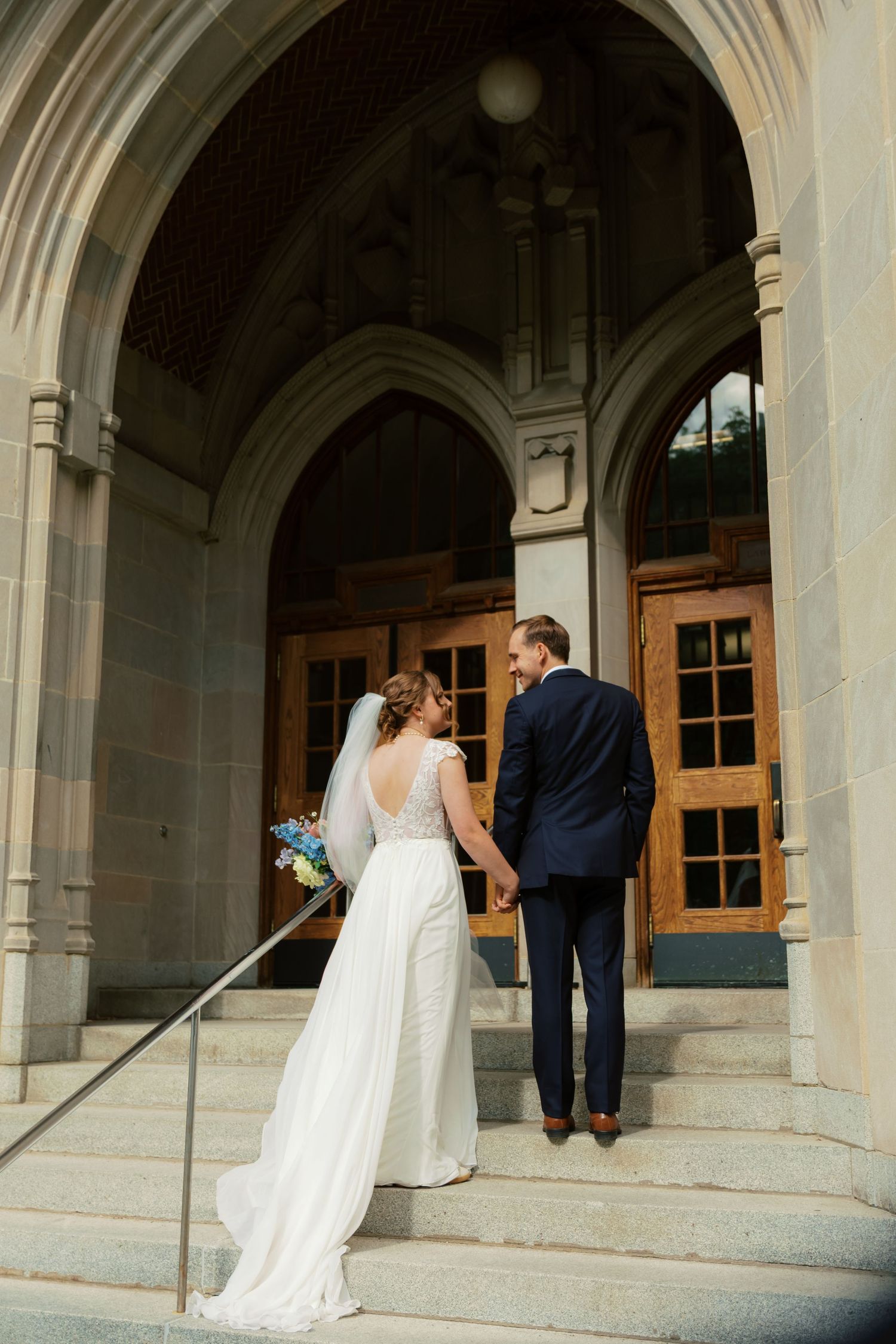 A bride and groom hold hands while walking up stone steps toward the entrance of a grand wedding venue.