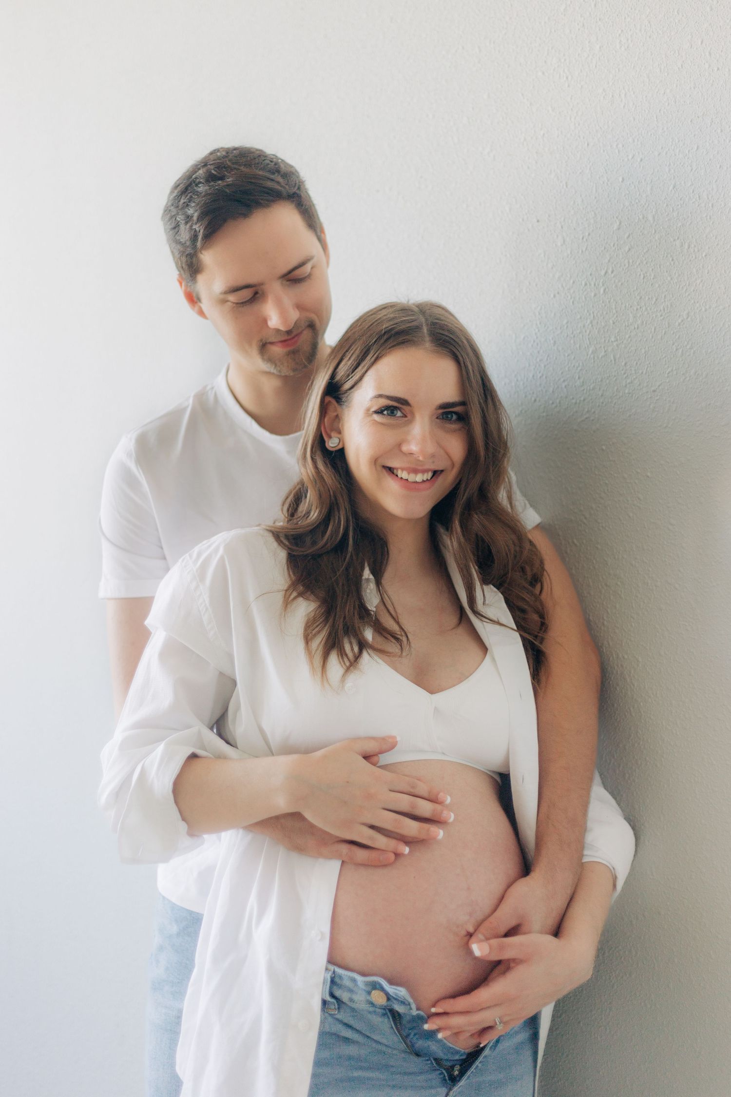 Expecting couple in white clothing embrace against a white wall during maternity photoshoot.