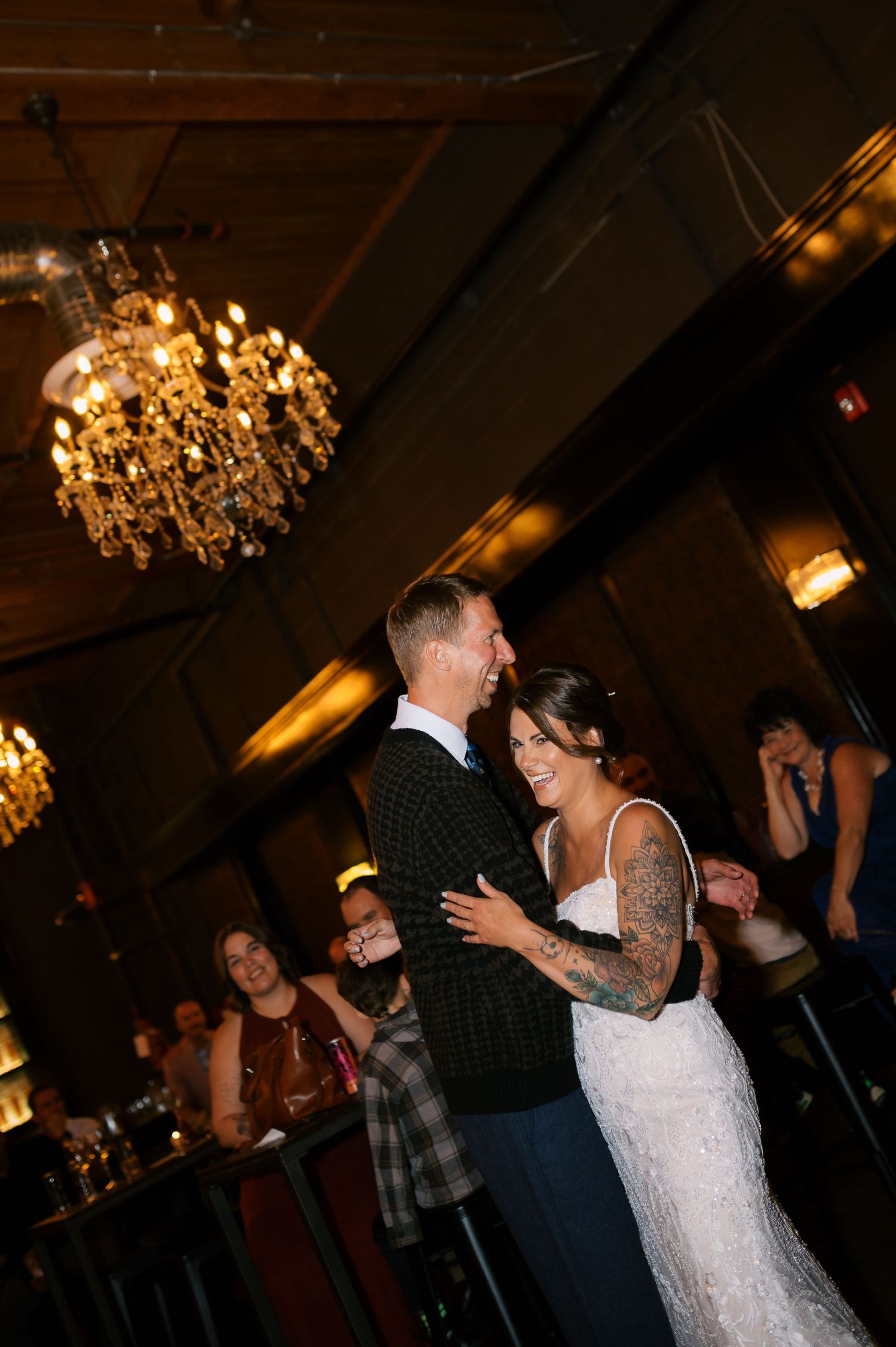 Couple sharing their first dance at a wedding reception under elegant chandeliers, bride in lace gown with tattoos.