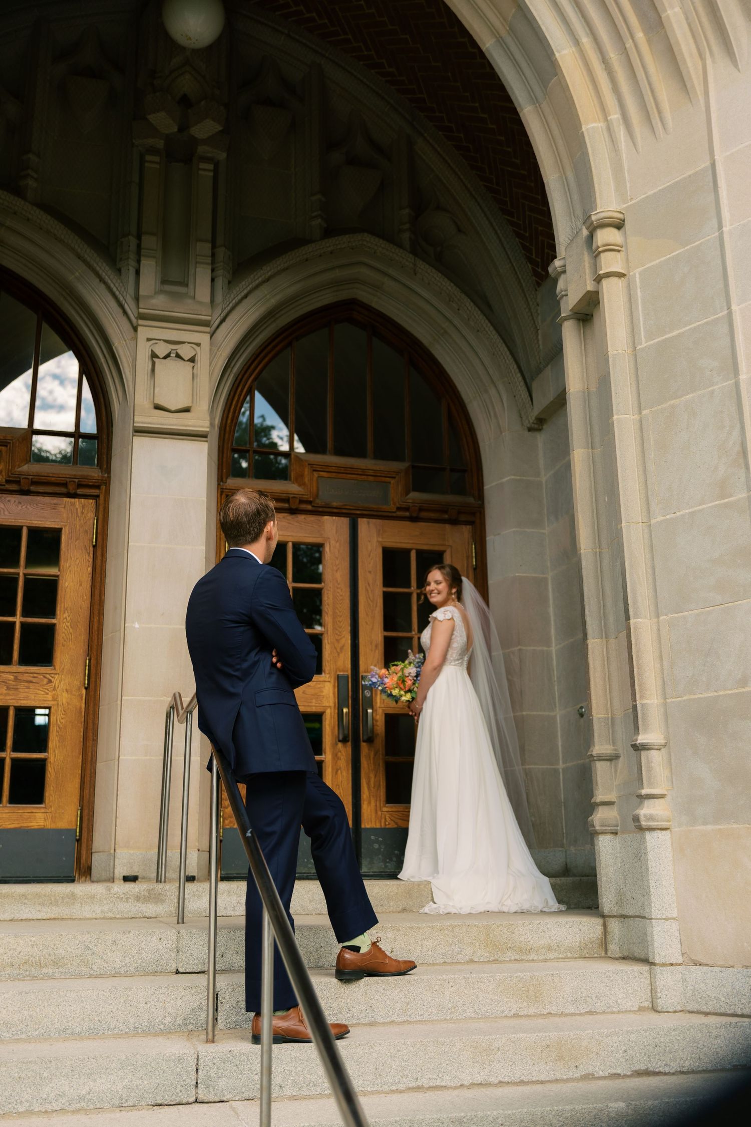 Bride in white gown and groom in navy suit share a first look on stone church steps with arched wooden doors.