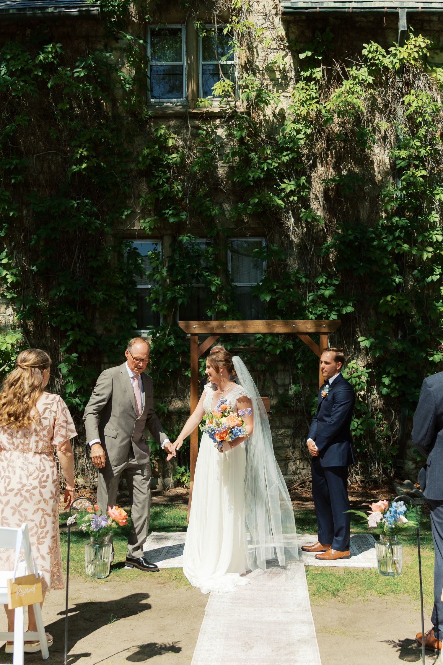 Bride walks down outdoor aisle with father, groom waiting at wooden arch, ivy-covered building backdrop.