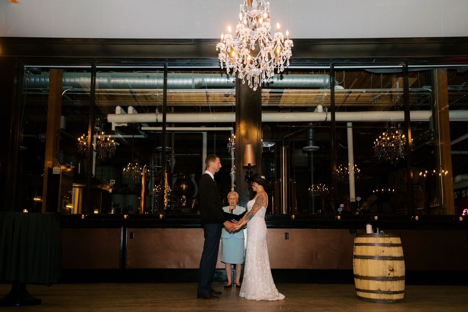 Bride and groom share first dance under elegant chandelier in rustic wedding venue with warm candlelit ambiance.