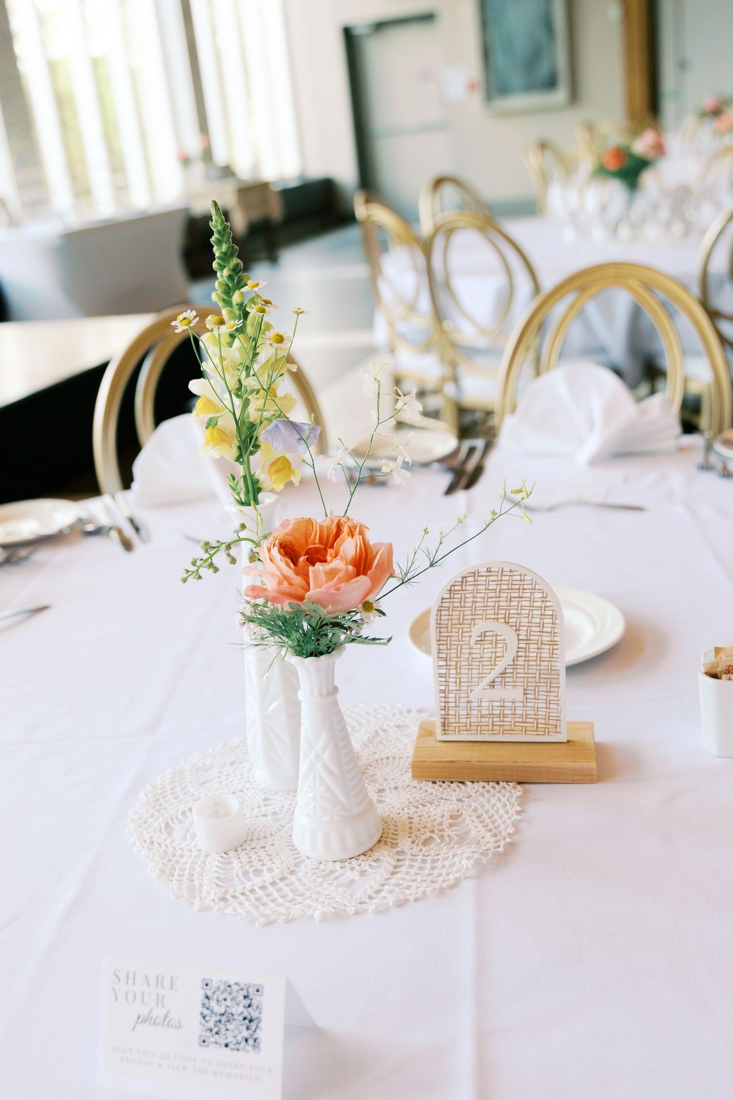 Elegant wedding table setting with white linen, milk glass bud vases, peach flowers, and table number 2.