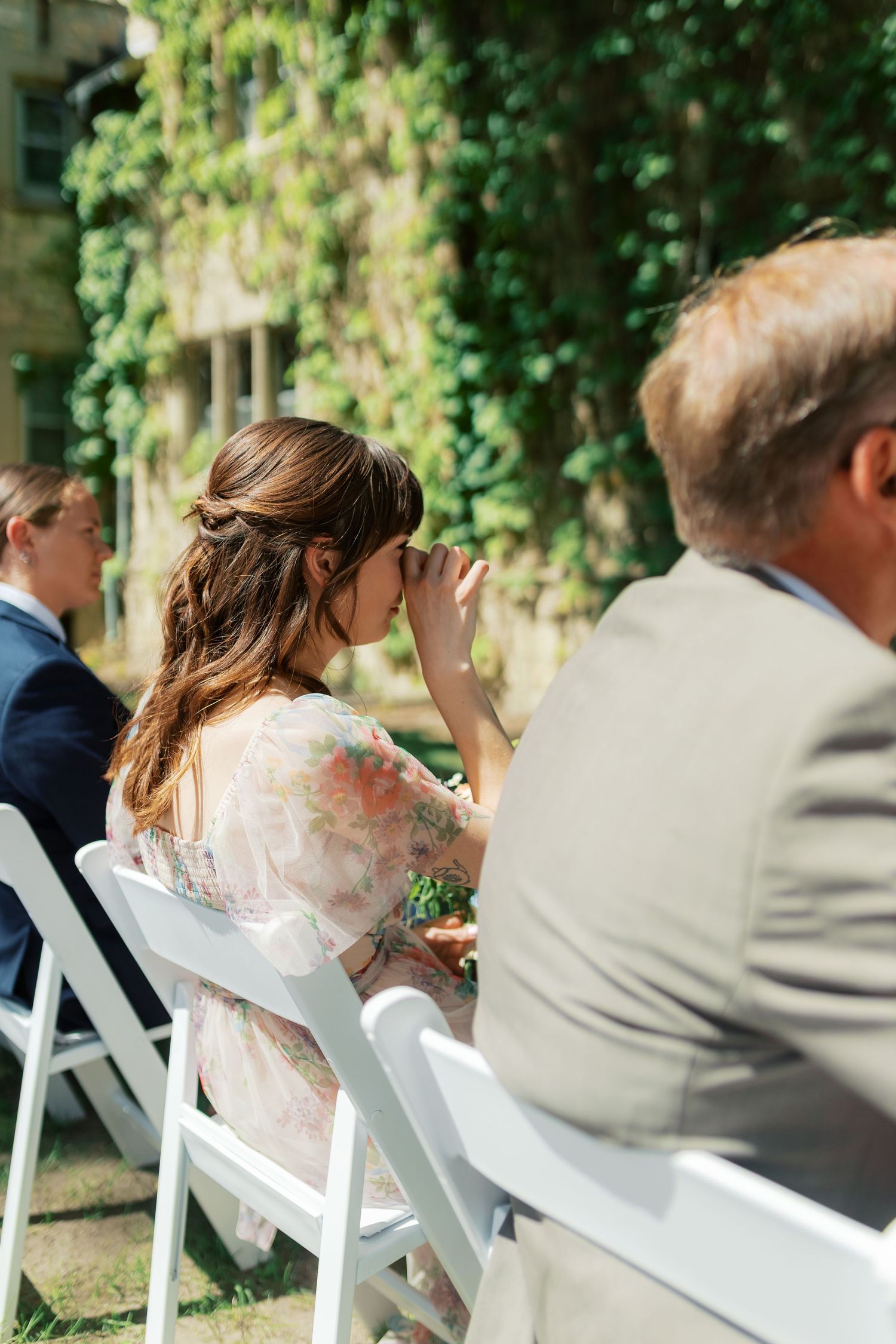 Emotional wedding guest in floral dress wiping tears during outdoor ceremony with ivy-covered building backdrop.