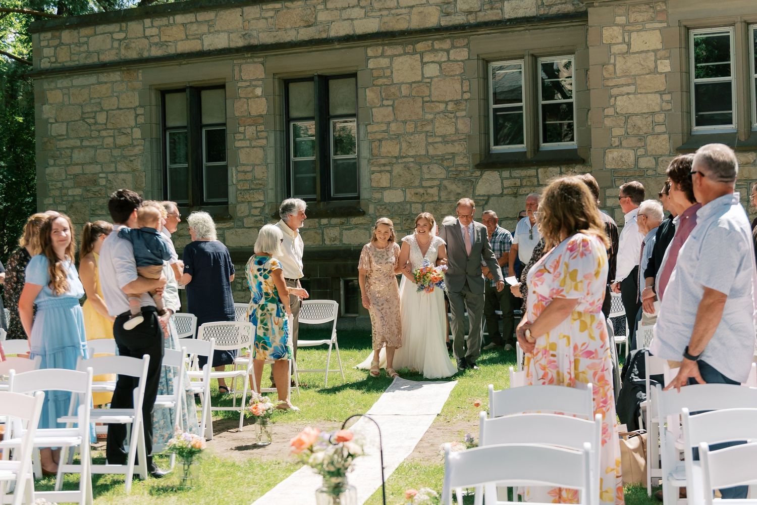Outdoor wedding ceremony with guests seated in white chairs on a sunny day at a stone building venue.