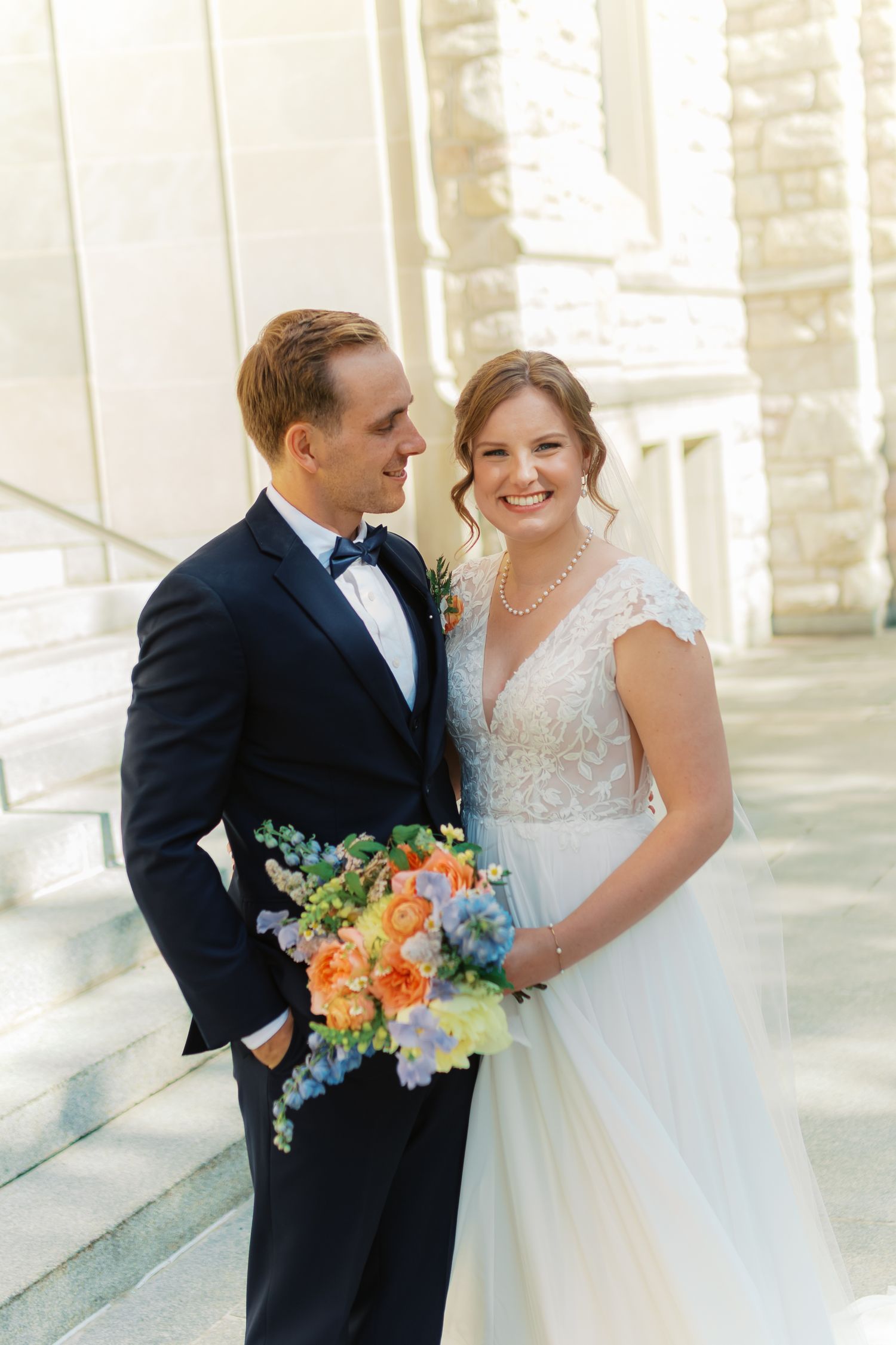 Smiling bride in lace gown holds colorful bouquet beside groom in navy suit at sunny outdoor wedding.