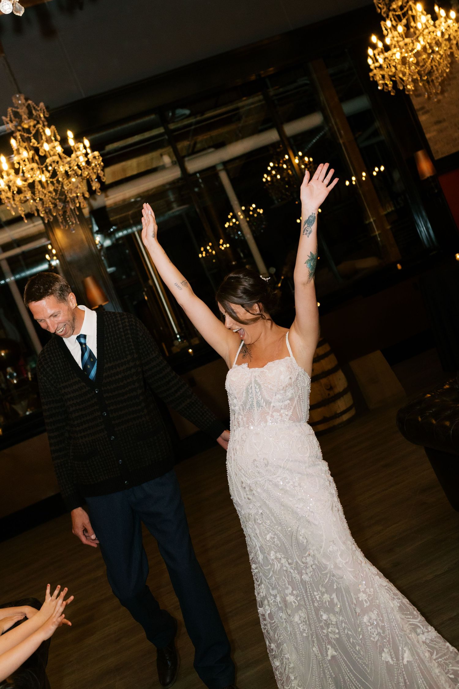 Bride in lace gown raises arms joyfully on dance floor with groom in navy suit at elegant chandelier-lit wedding reception.