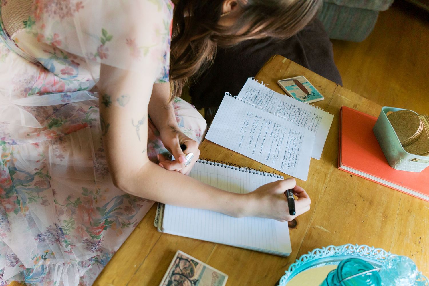 Young woman in floral dress writing in spiral notebook at wooden desk with school supplies.