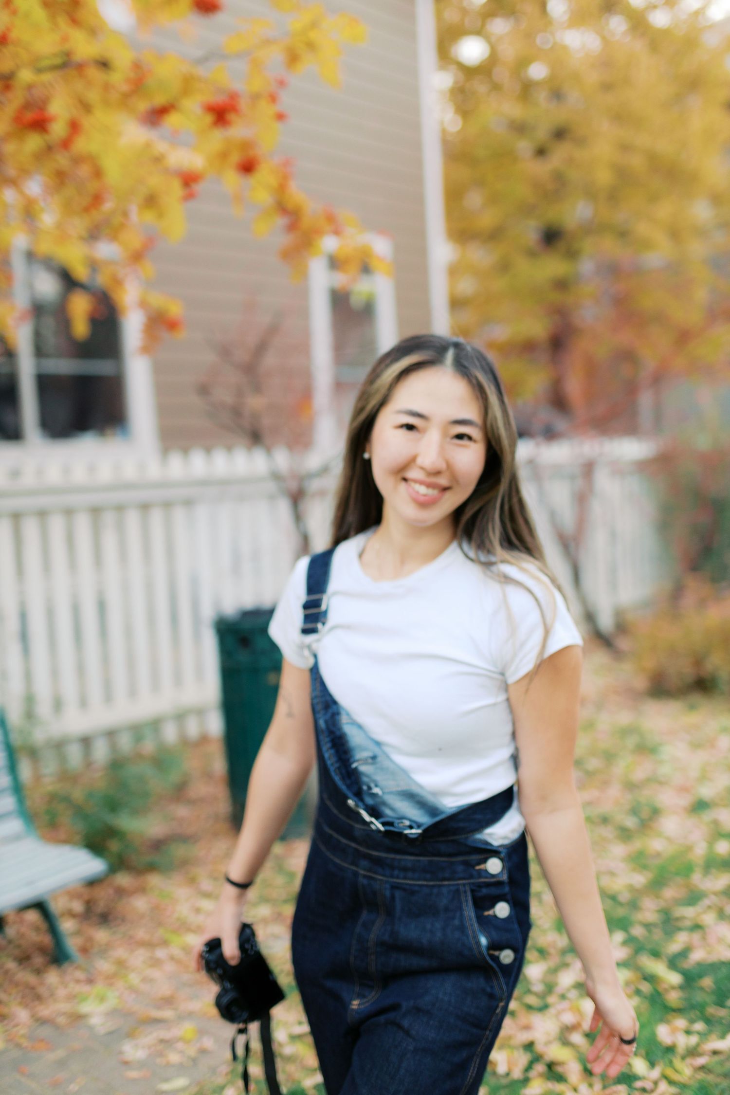 Woman in denim overalls smiling on autumn porch with fall foliage in background.