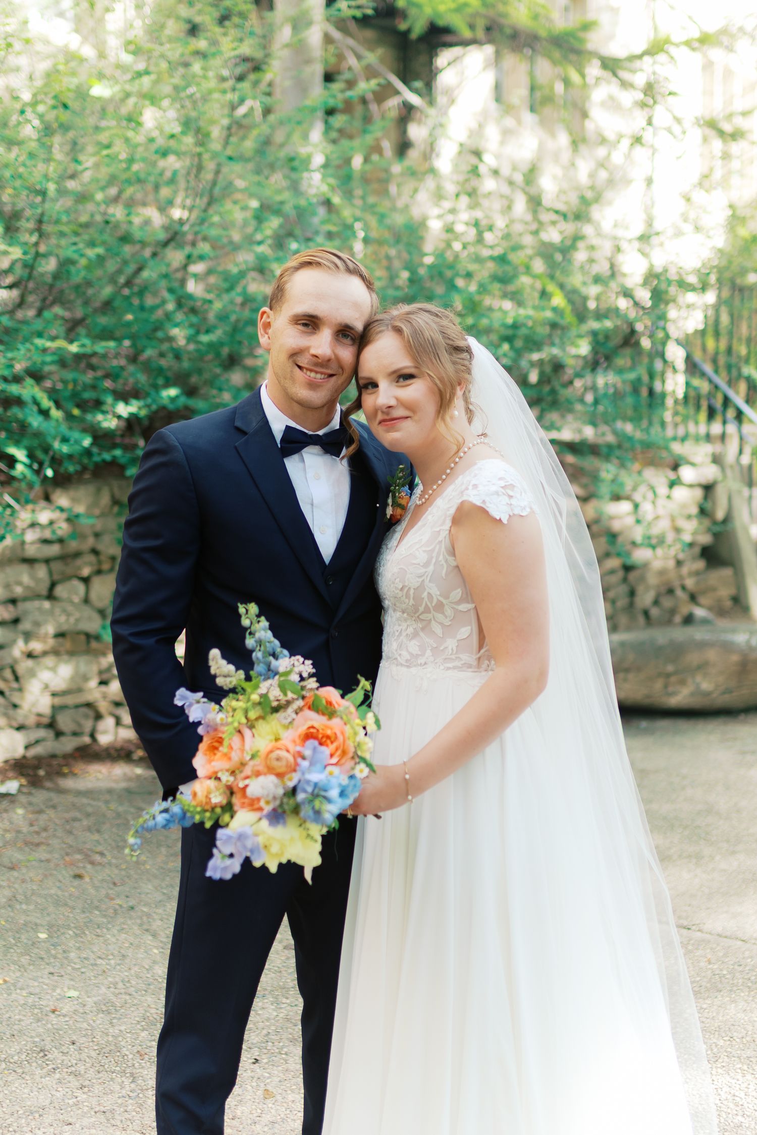 Bride and groom pose together outdoors, she holds a colorful bouquet of orange and blue flowers, wearing a lace gown and veil.