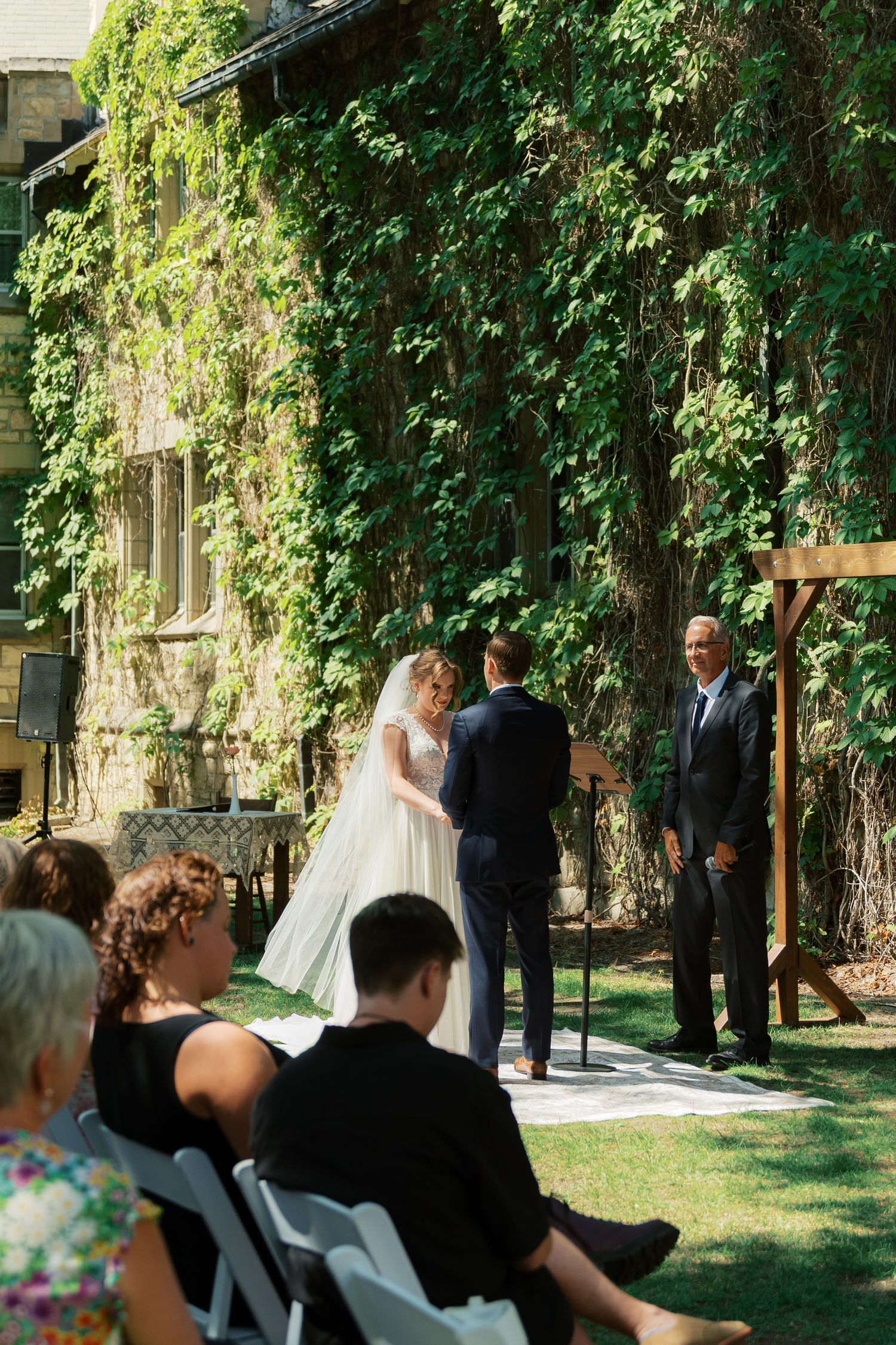 Outdoor wedding ceremony with bride and groom exchanging vows on green lawn before ivy-covered stone building.