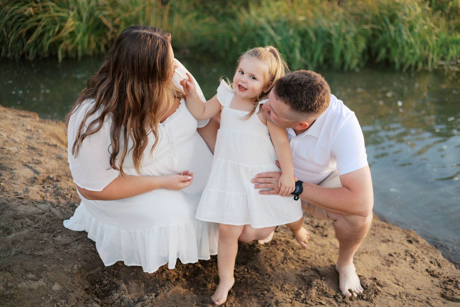 A couple in white outfits share a joyful moment with a young girl by a waterfront at sunset.