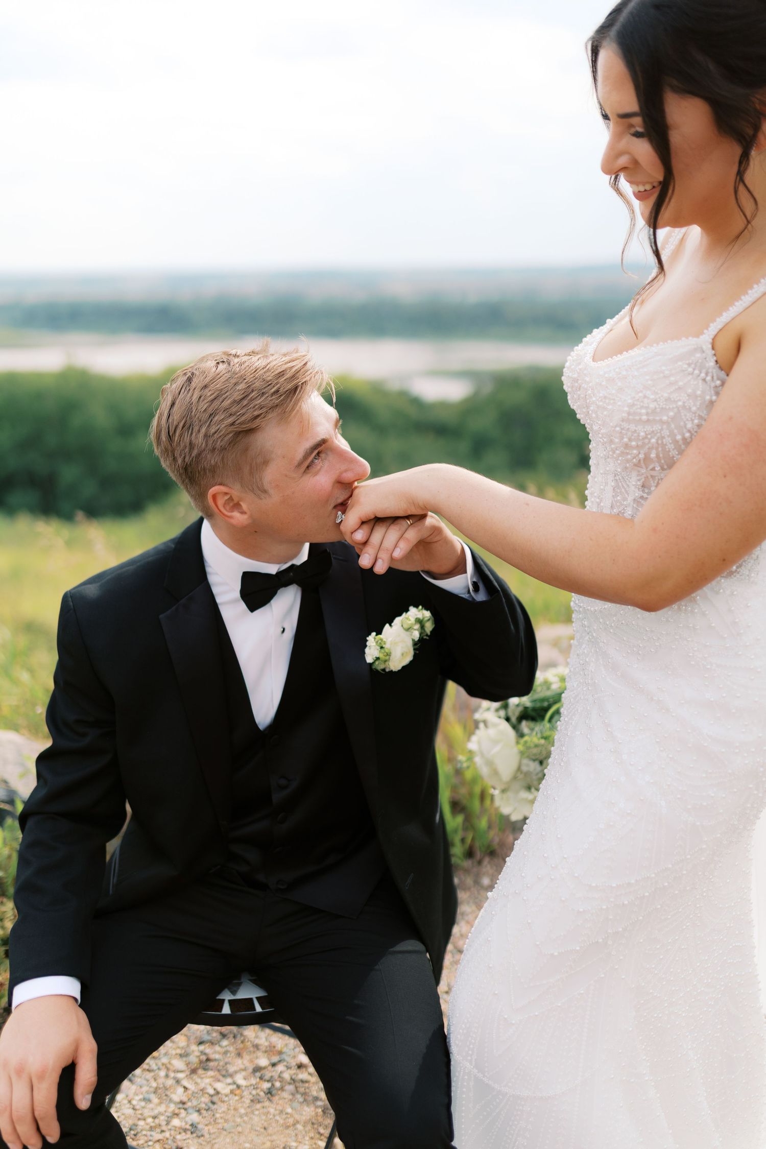 Groom in black tuxedo kisses bride's hand while she smiles, outdoors with greenery background.