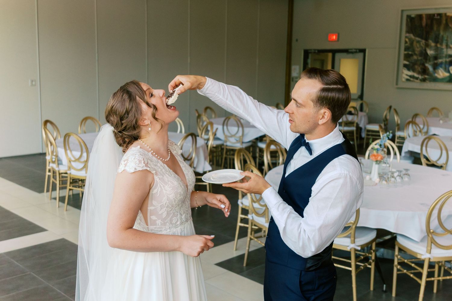 Groom feeding bride a bite of food at wedding reception with gold chairs and white tablecloths in background.