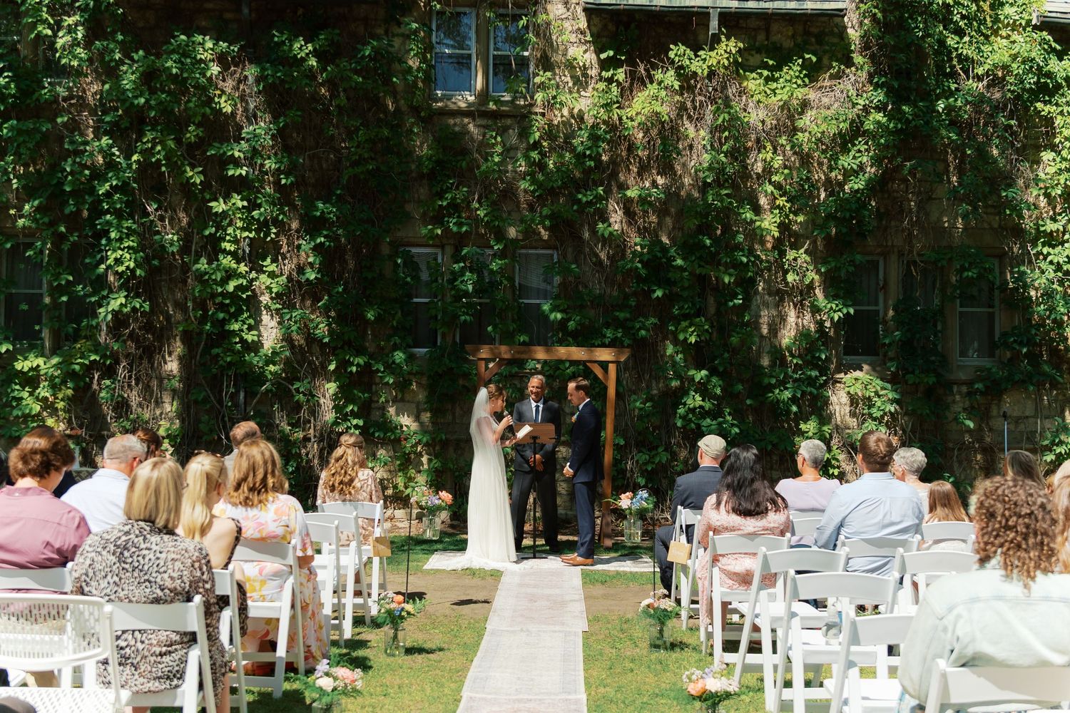 Outdoor wedding ceremony with bride and groom at floral arch, guests seated on white chairs, ivy-covered building backdrop.