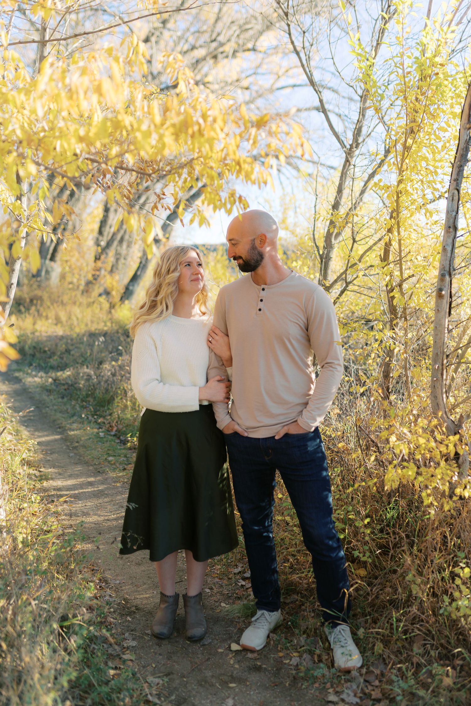 Couple standing on a autumn forest path, woman in white sweater and green skirt, man in beige shirt and jeans.
