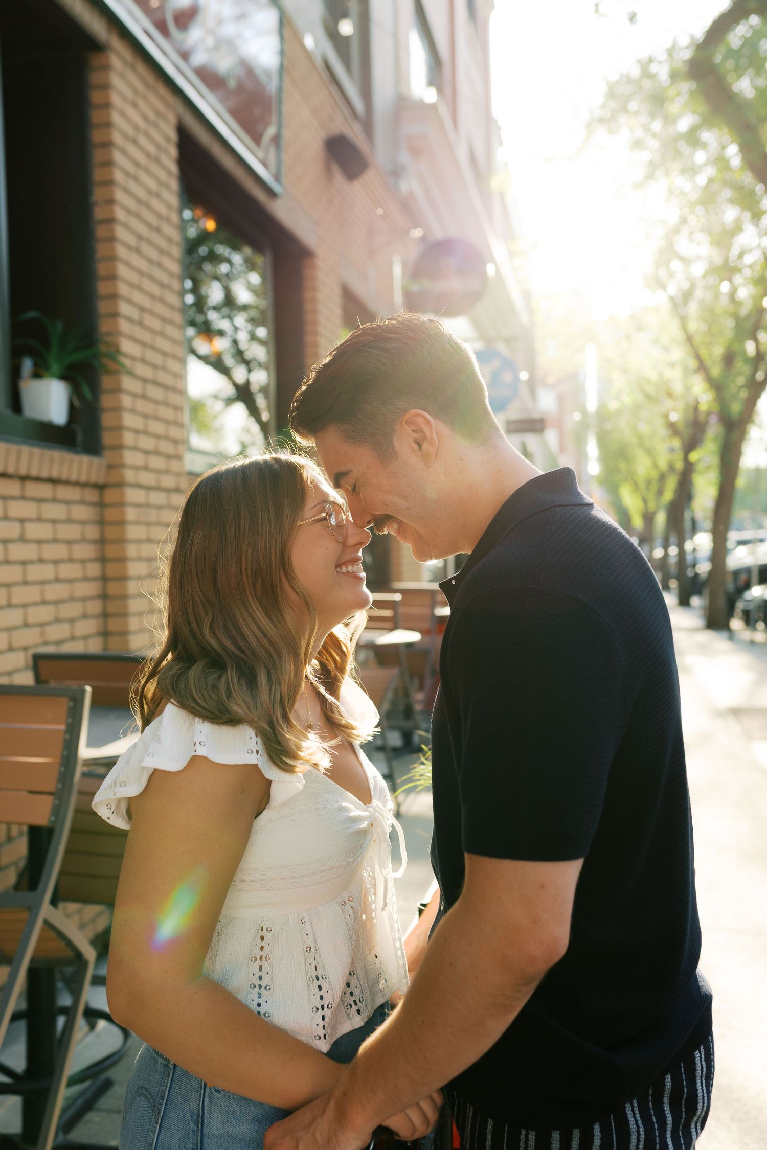 A romantic couple shares an intimate moment on a sun-lit sidewalk outside a brick building with cafe seating.